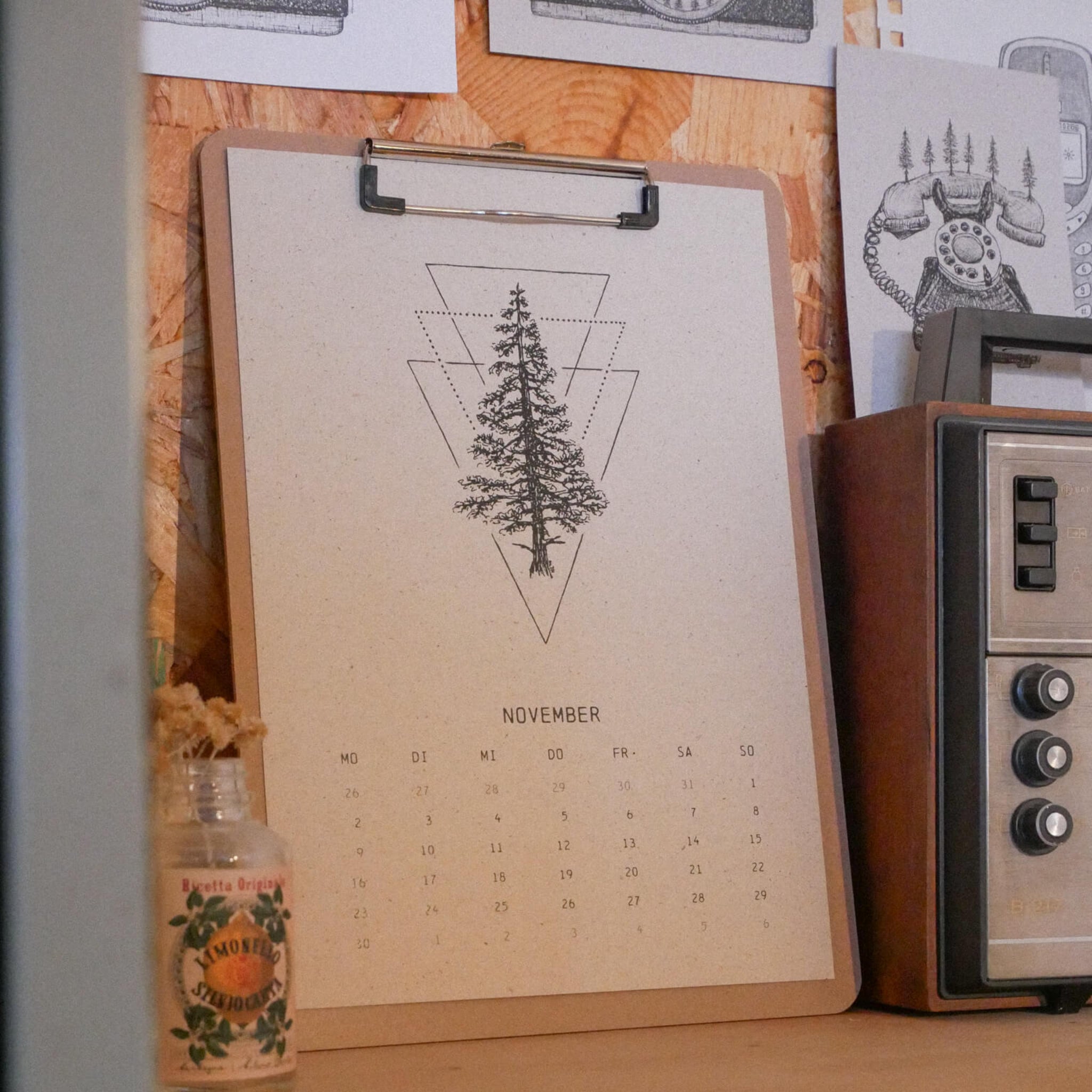 Ein Klemmbrett mit einem minimalistischen November-Kalender mit einer Tannenbaum-Illustration liegt auf einer Holzfläche aus. In der Nähe stehen ein altes Radio, eine kleine dekorative Flasche und schwarz-weiße Kunstdrucke an der Wand.
