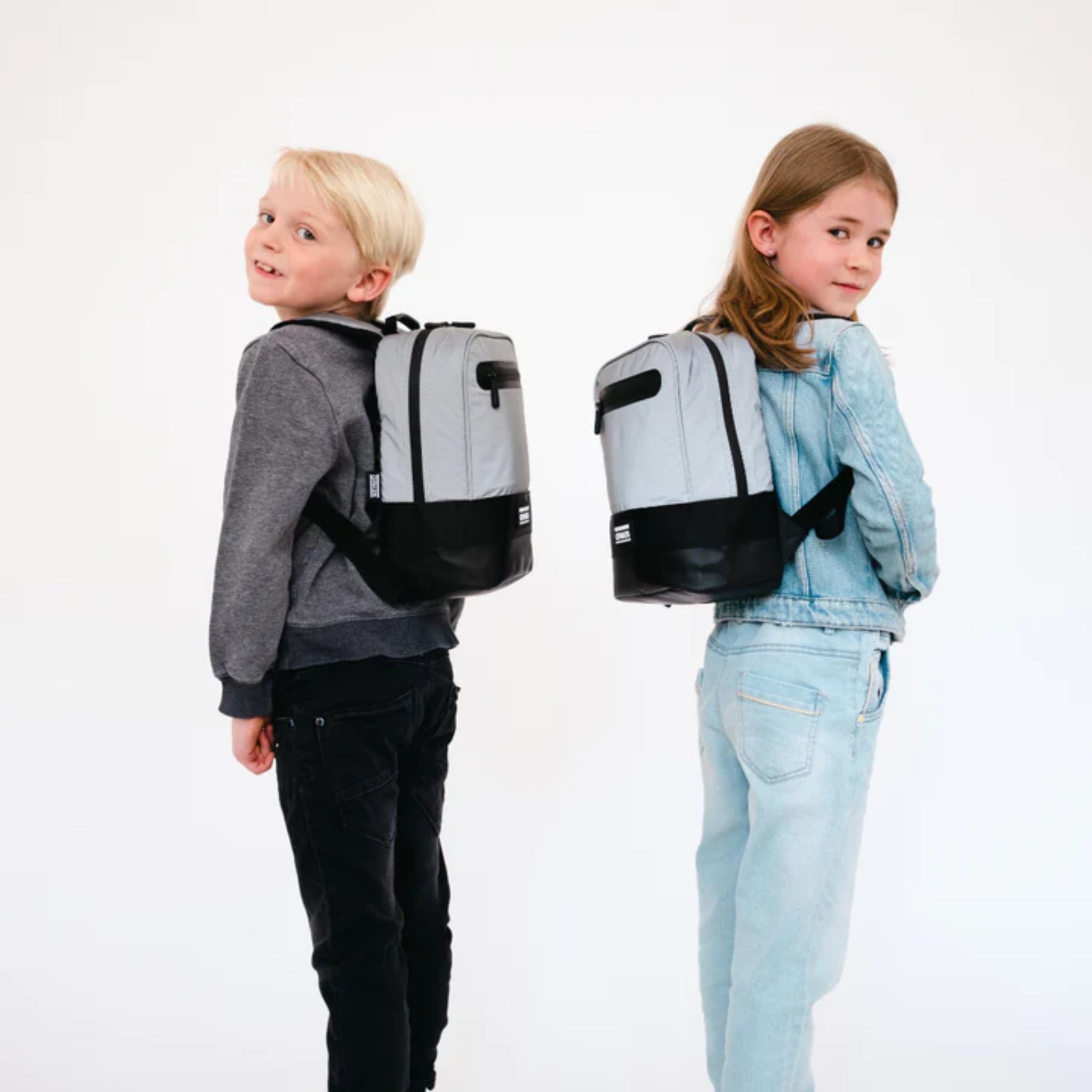 Zwei Kinder tragen graue Rucksäcke und stehen Rücken an Rücken vor einem weißen Hintergrund.