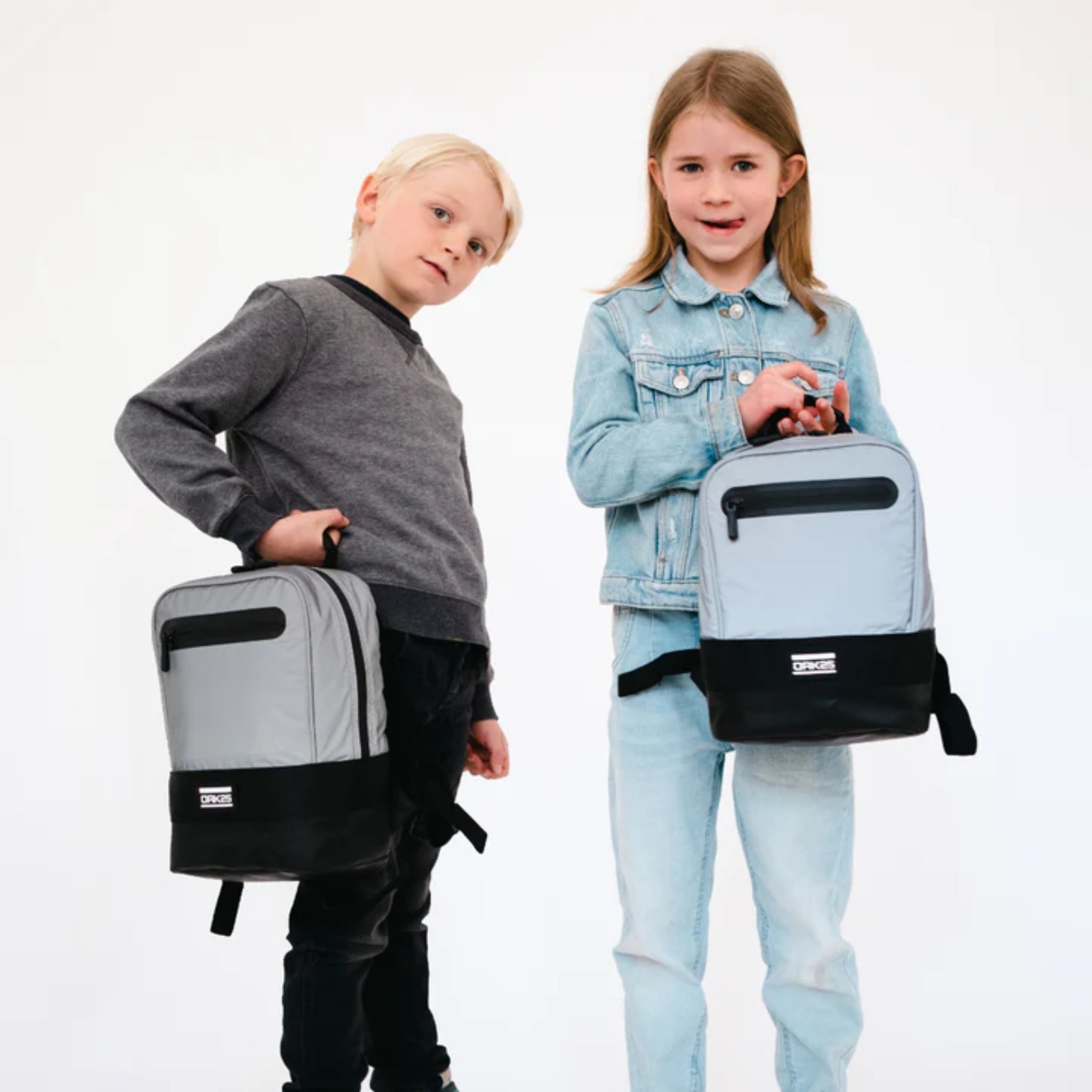 Zwei Kinder posieren mit grauen Rucksäcken, das Mädchen trägt eine Jeansjacke, der Junge einen grauen Pullover.