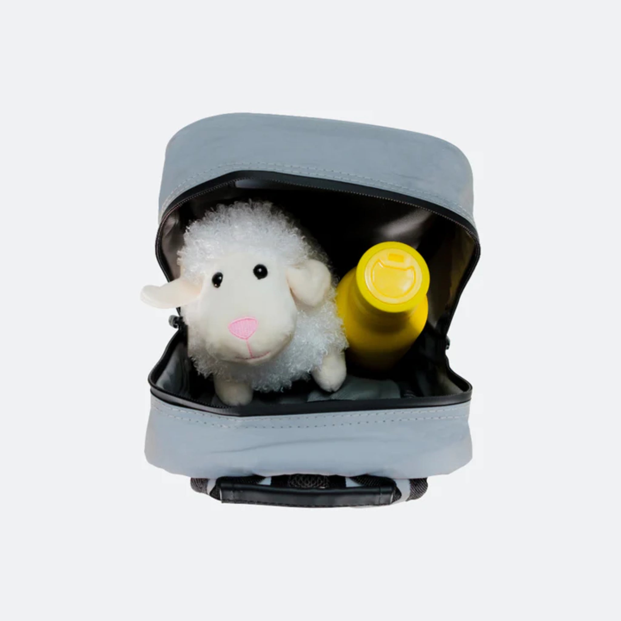Graue Tasche mit offenem Reißverschluss, darin ein Plüschschaf und eine gelbe Flasche vor weißem Hintergrund.
