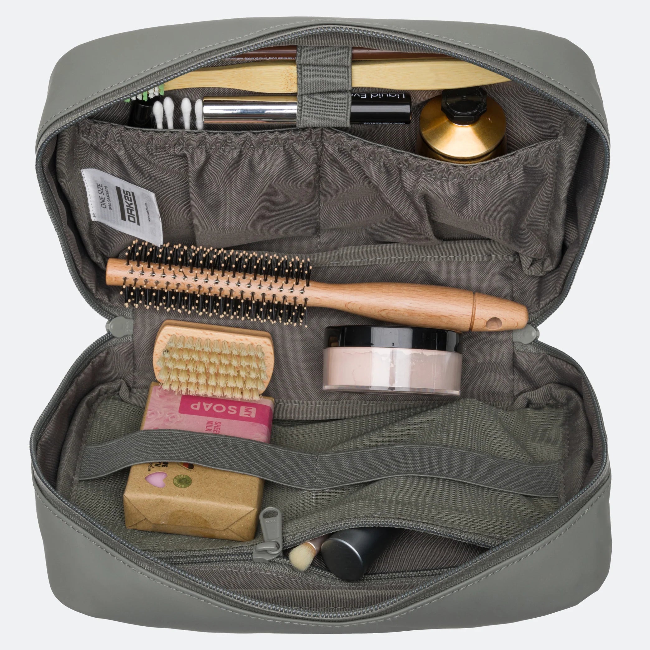 Ein grauer Kulturbeutel wird geöffnet, und es kommen Gegenstände zum Vorschein, darunter eine runde Haarbürste, ein Stück Seife, ein Cremetiegel, eine kleine Flasche, Wattestäbchen, eine Zahnbürste, Zahnpasta und eine Schminkbürste.