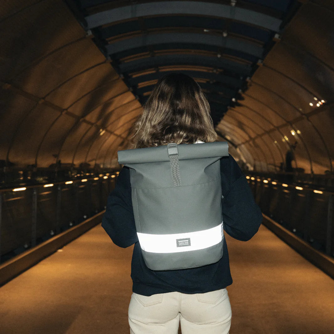 Eine Person mit schulterlangen Haaren, die eine dunkle Jacke und eine beigefarbene Hose trägt, steht mit dem Rücken zur Kamera in einem Tunnel und trägt einen grauen Rucksack mit einem reflektierenden Streifen.