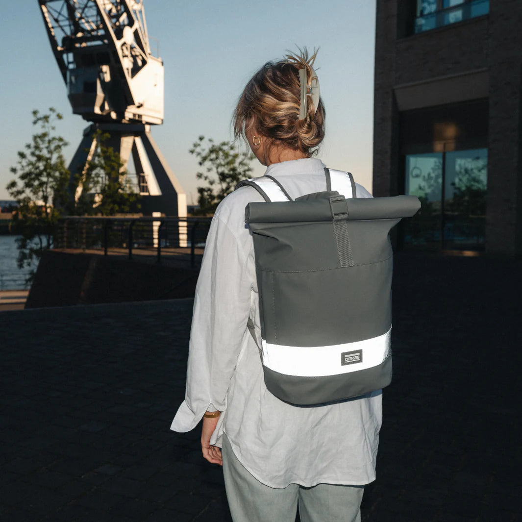 Eine Person mit hellen Haaren in einer Spange, die ein helles Hemd und einen großen grauen Rolltop-Rucksack mit einem reflektierenden weißen Streifen trägt, steht im Freien in der Nähe eines Industriekrans und eines Gebäudes im Abendlicht.