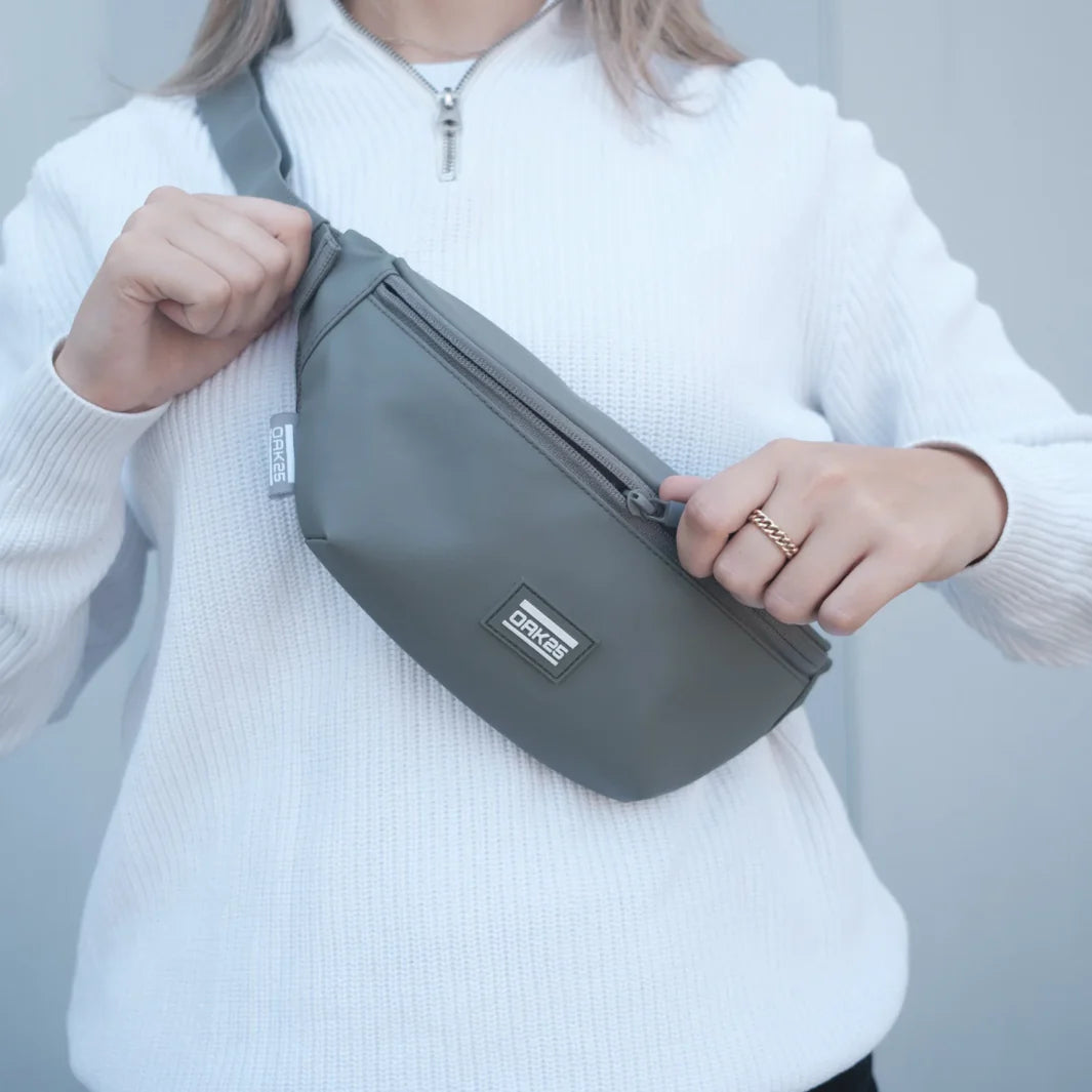 Eine Person, die einen weißen Pullover mit Reißverschluss trägt, hält eine graue ORKEE-Umhängetasche und öffnet den Reißverschluss. Nur der Oberkörper und die Hände sind vor einem einfarbigen Hintergrund zu sehen.