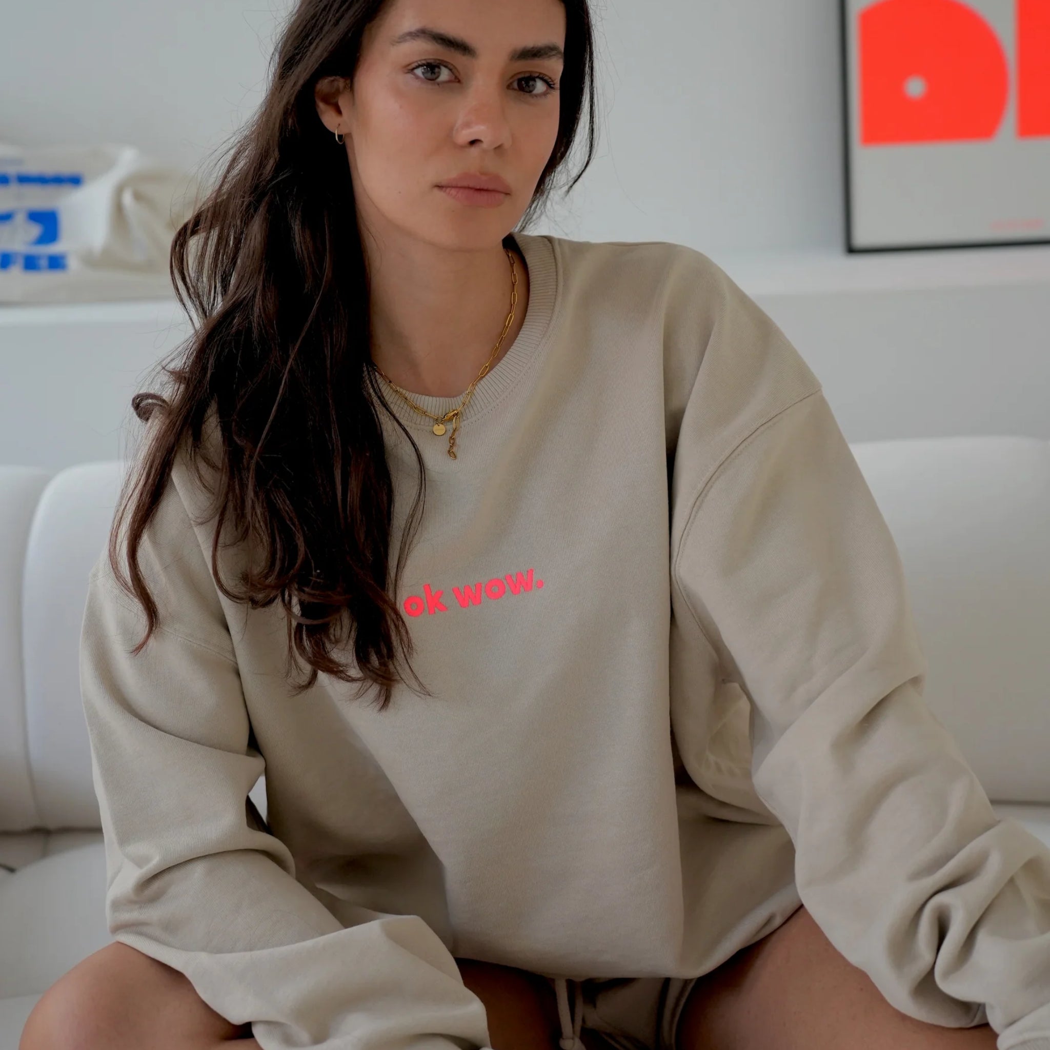 Eine Frau mit langen braunen Haaren sitzt auf einer weißen Couch und trägt ein beiges Sweatshirt in Übergröße mit der Aufschrift ok wow. in rosa Schrift. Sie blickt mit einem neutralen Blick in die Kamera. Im Hintergrund ist eine minimalistische Einrichtung zu sehen.