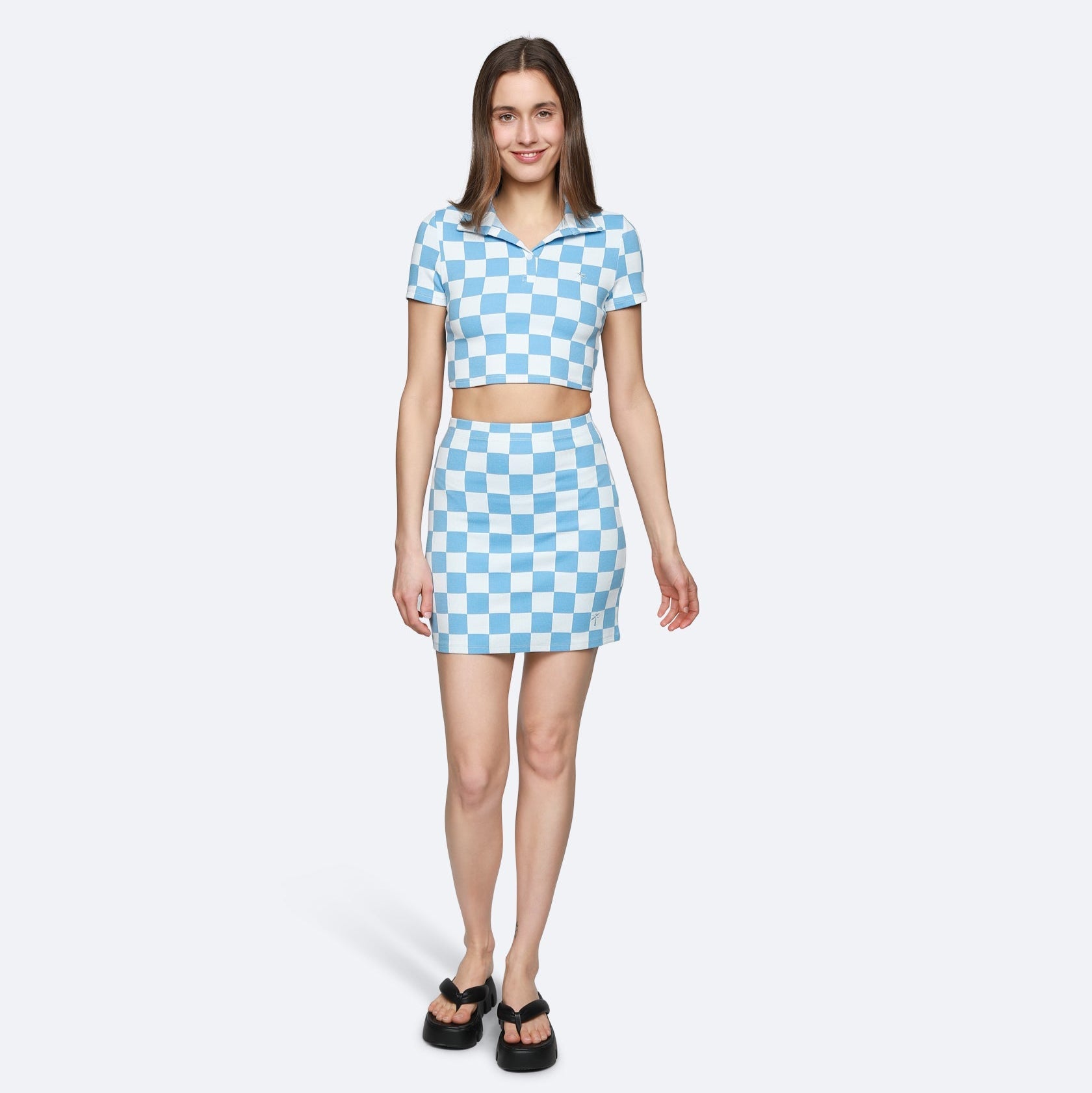 Eine Frau steht lächelnd da und trägt ein blau-weiß kariertes, kurzärmeliges Crop-Top und einen dazu passenden Rock mit hoher Taille, gepaart mit schwarzen Sandalen. Sie posiert vor einem schlichten hellen Hintergrund.