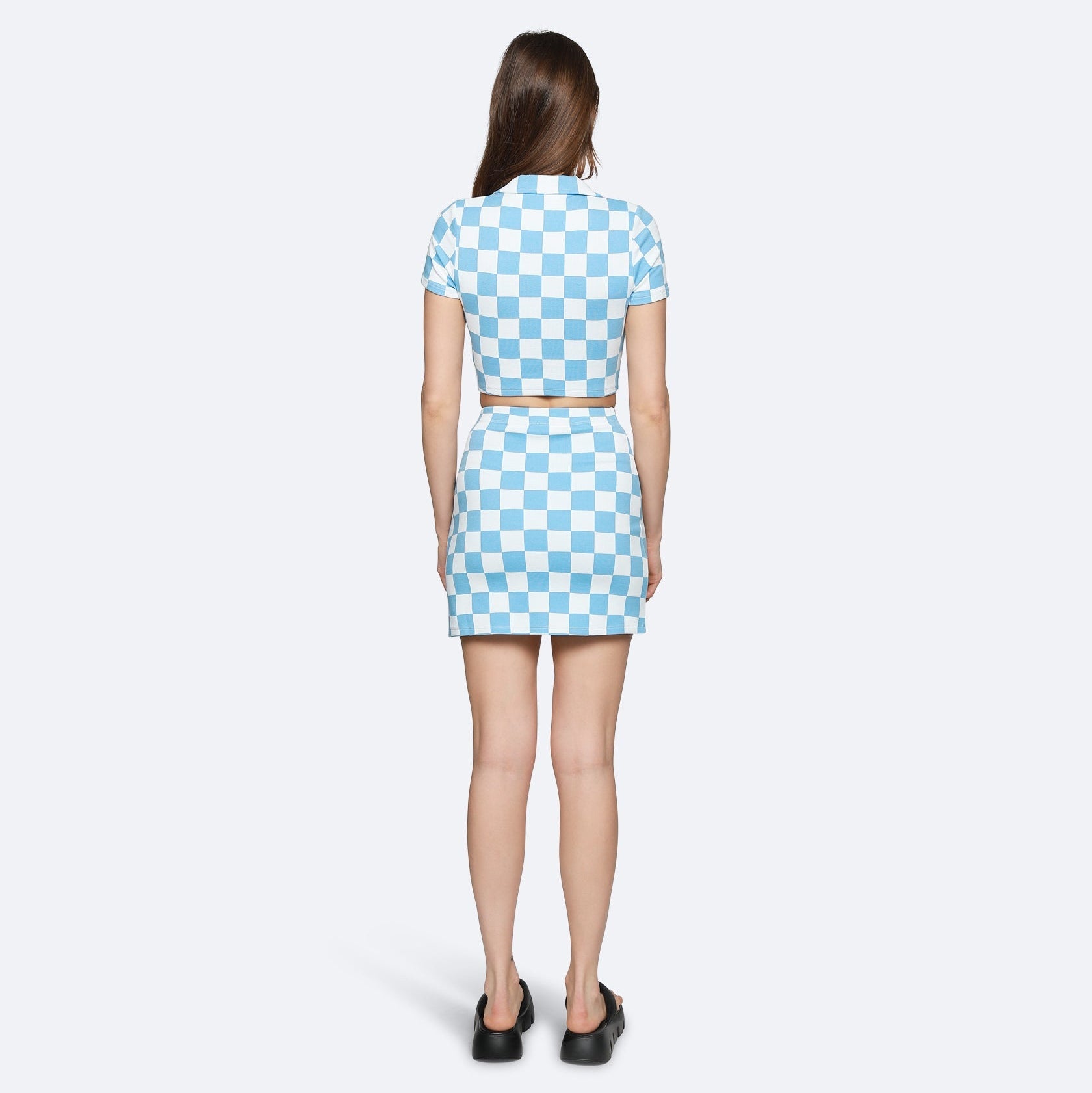 Eine Frau steht mit dem Gesicht zur Wand und trägt ein kurzärmeliges, blau-weißes, schachbrettartig gemustertes Kleid mit einem beigen Gürtel und schwarzen Slippern vor einem schlichten, hellen Hintergrund.