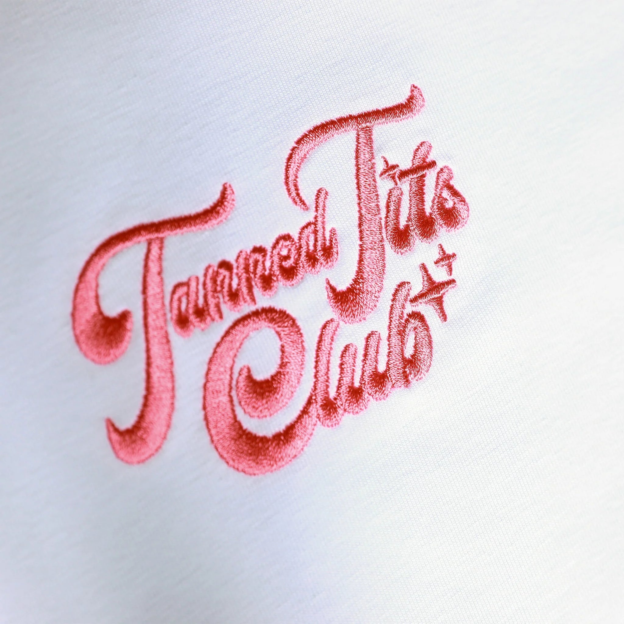 Close-up eines Stoffes mit roter, gestickter Schrift, die "Tanned Tits Club" in verspielter Schrift zeigt.