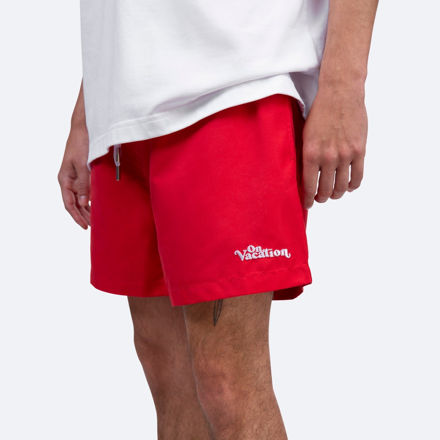 Eine Person, die ein weißes T-Shirt und leuchtend rote Shorts mit weißer Stickerei "On Vacation" auf dem linken Bein trägt, steht vor einem schlichten hellen Hintergrund.