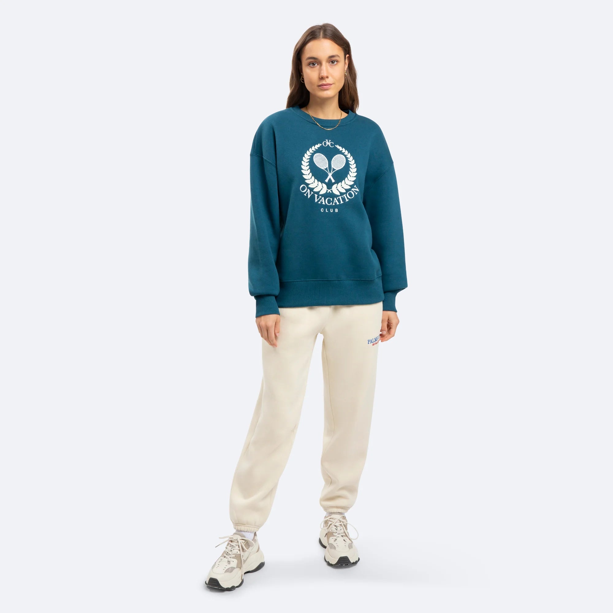 Frau in blauem Pullover mit der Aufschrift "ON VACATION" und beiger Jogginghose steht vor neutralem Hintergrund.