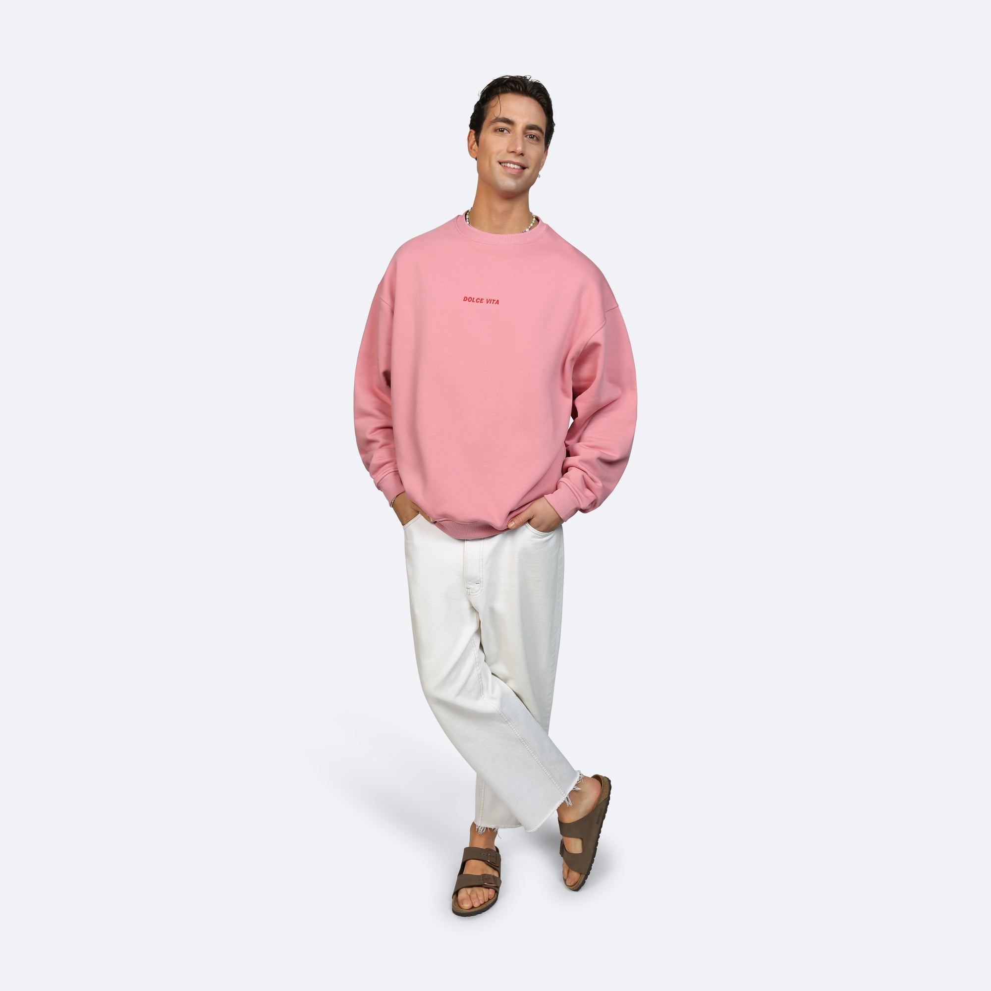 Eine Person steht lächelnd mit den Händen in den Taschen, trägt ein rosa Sweatshirt in Übergröße, weiße Hosen und braune Sandalen vor einem schlichten hellgrauen Hintergrund.
