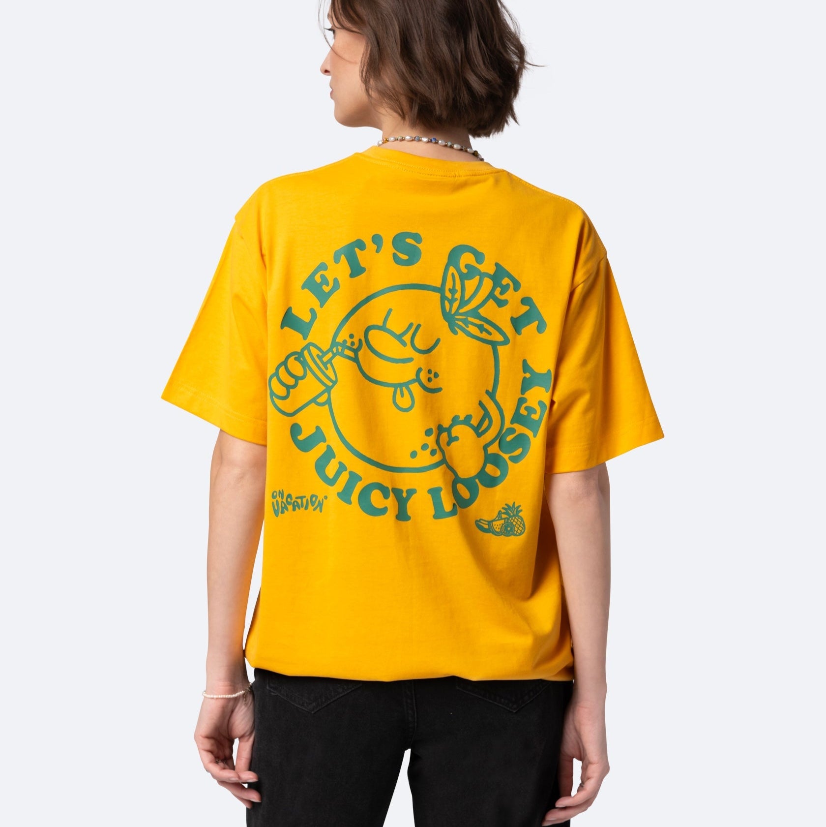 Person, die ein leuchtend gelbes T-Shirt mit einer großen orangefarbenen Karikatur und dem Text LETS GET JUICY LOOSEY auf dem Rücken in grüner Farbe trägt. Die Person blickt von der Kamera weg.