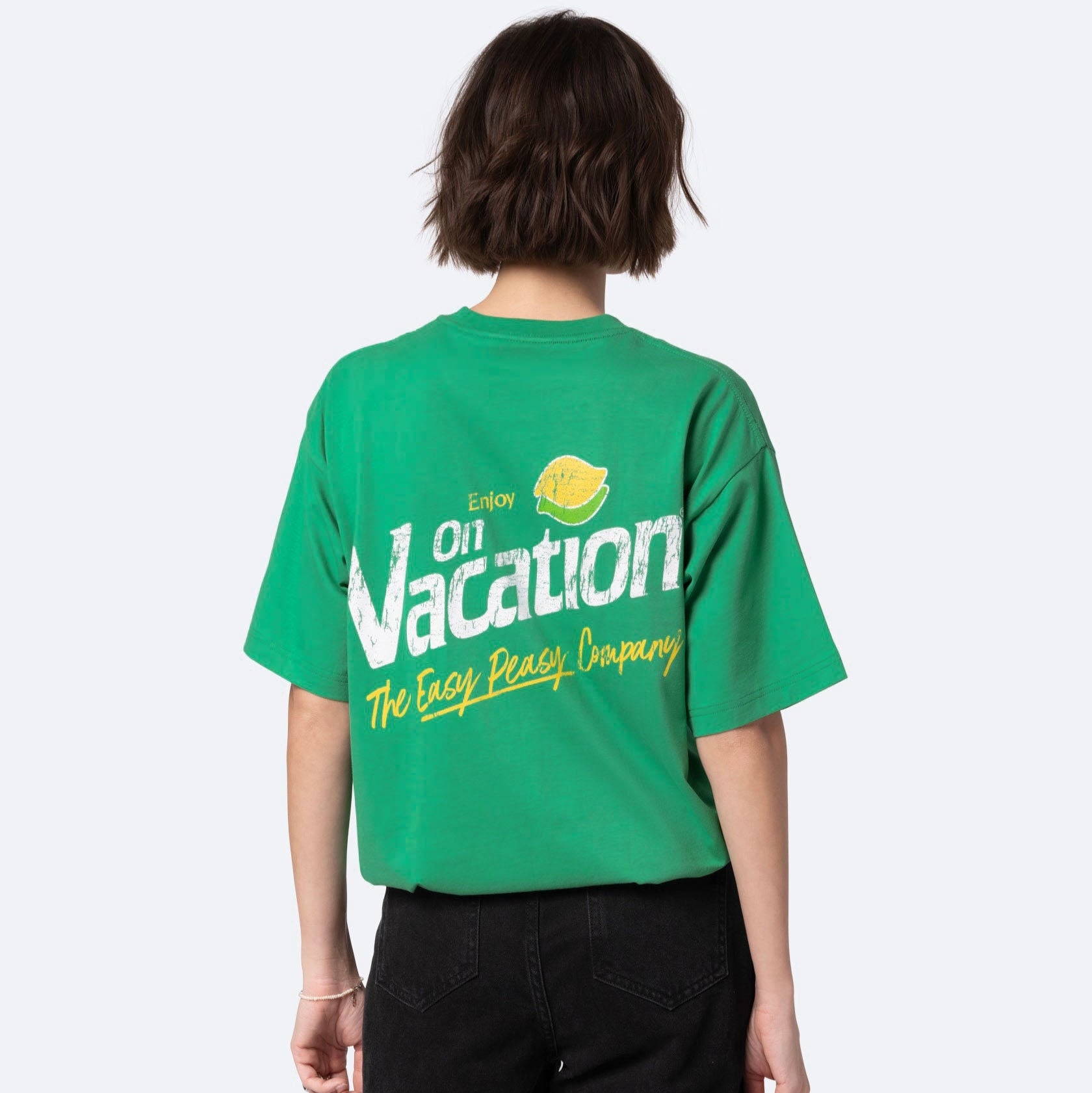 Eine Person mit kurzen braunen Haaren trägt ein grünes T-Shirt mit der Aufschrift Enjoy On Vacation The Easy Peasy Company in fetten weißen und gelben Buchstaben, dazu eine schwarze Jeans. Die Person blickt von der Kamera weg.
