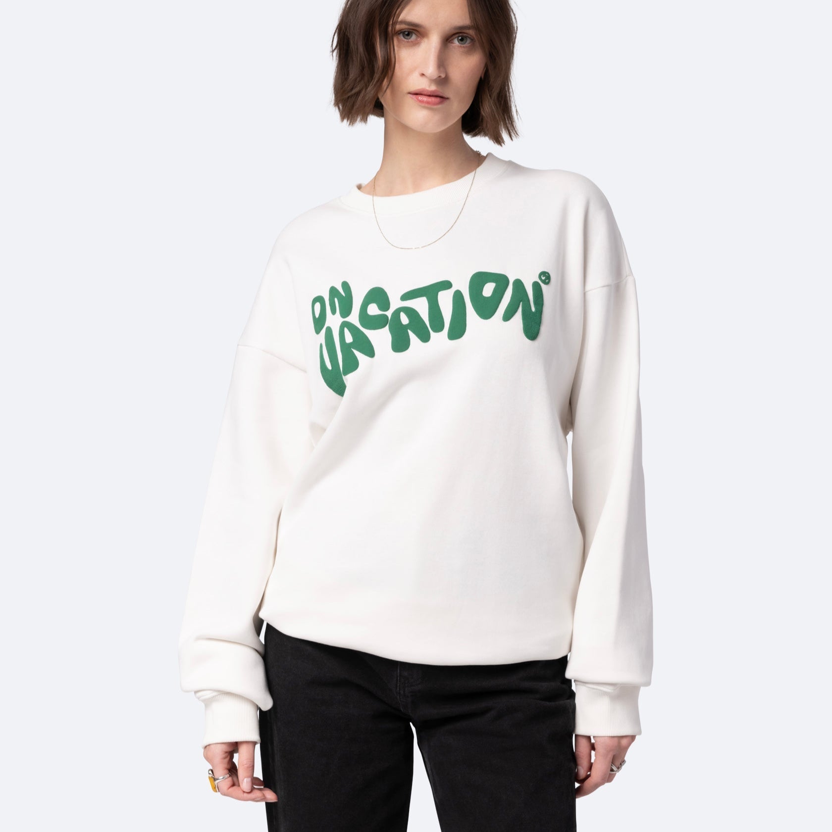 Eine Person mit kurzen braunen Haaren trägt ein weißes Sweatshirt mit der Aufschrift "ON VACATION" in grüner Sprechblasenschrift und eine schwarze Hose vor einem schlichten hellgrauen Hintergrund.
