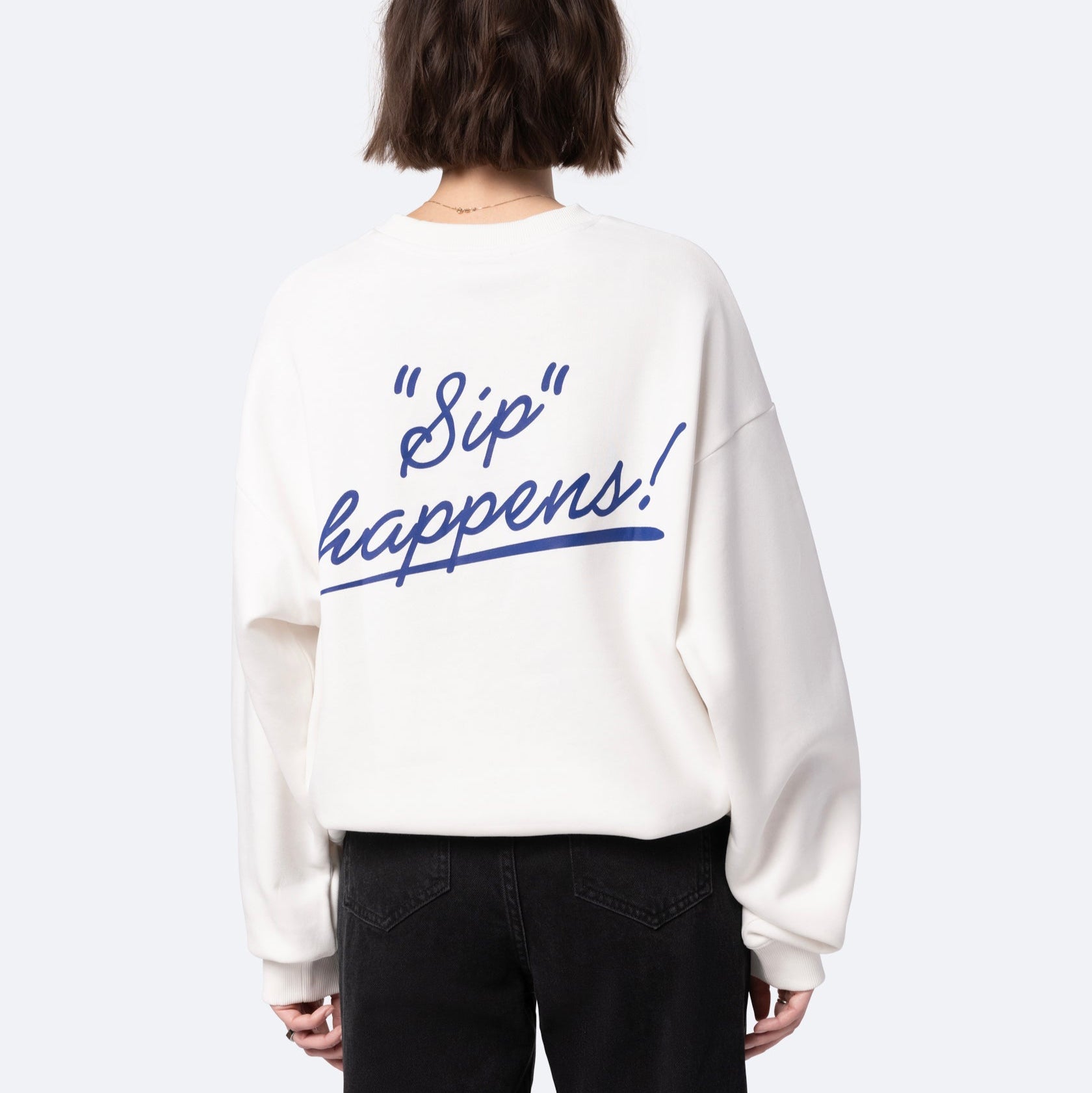 Eine Person mit kurzen braunen Haaren, von hinten gesehen, trägt ein weißes Sweatshirt mit der Aufschrift "Sip happens!" in blauer Schreibschrift auf dem Rücken. Außerdem trägt sie eine schwarze Jeans.
