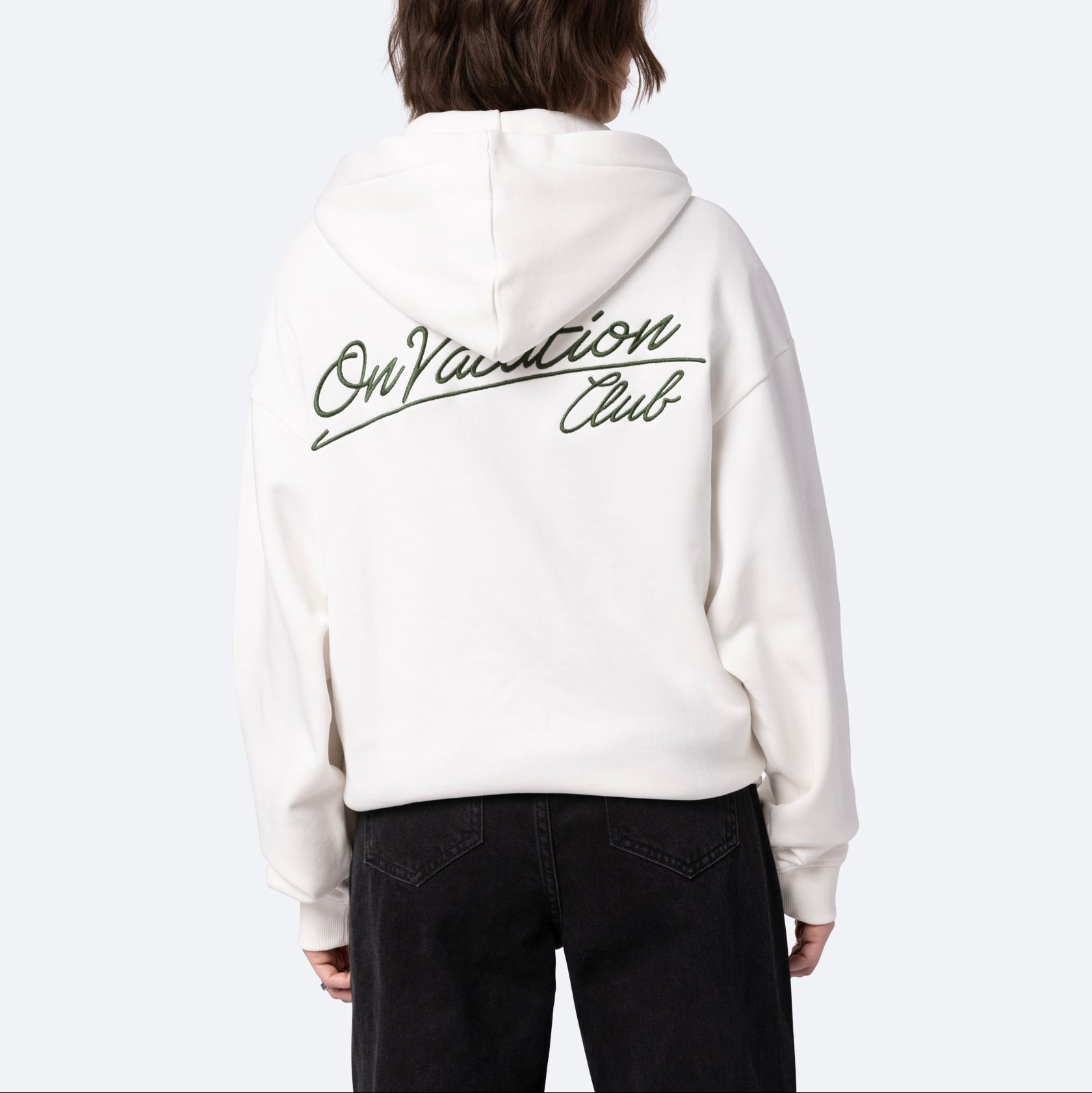 Eine Person mit schulterlangen braunen Haaren, von hinten gesehen, trägt einen weißen Kapuzenpullover mit dem Schriftzug On Vacation Club in grüner Schrift auf dem Rücken, dazu eine schwarze Hose.