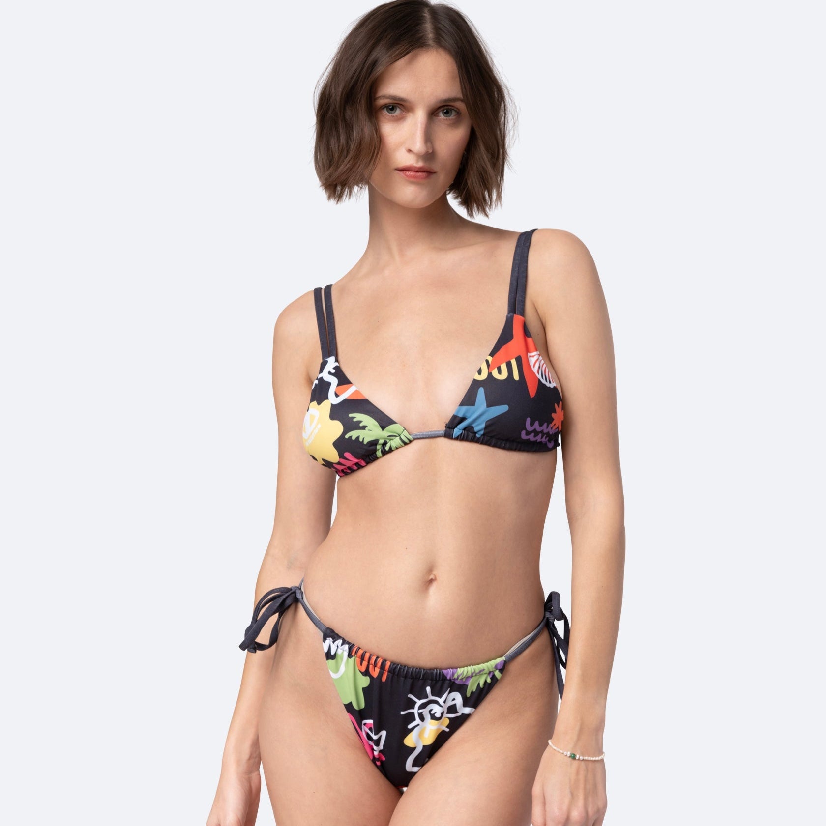 Eine Frau mit kurzen braunen Haaren trägt einen farbenfrohen Bikini mit Cartoon-Aufdruck und seitlich gebundener Hose und steht vor einem schlichten hellen Hintergrund.
