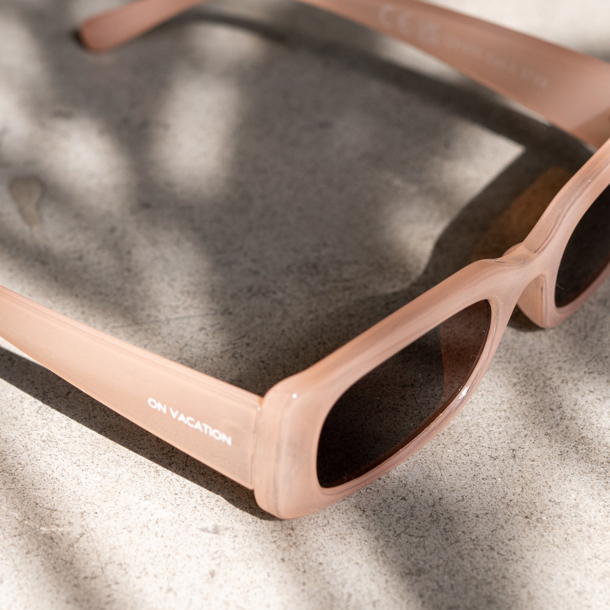 Beige Sonnenbrille mit der Aufschrift „On Vacation“ auf einem hellen Steinboden, beschattet von Sonnenlicht.