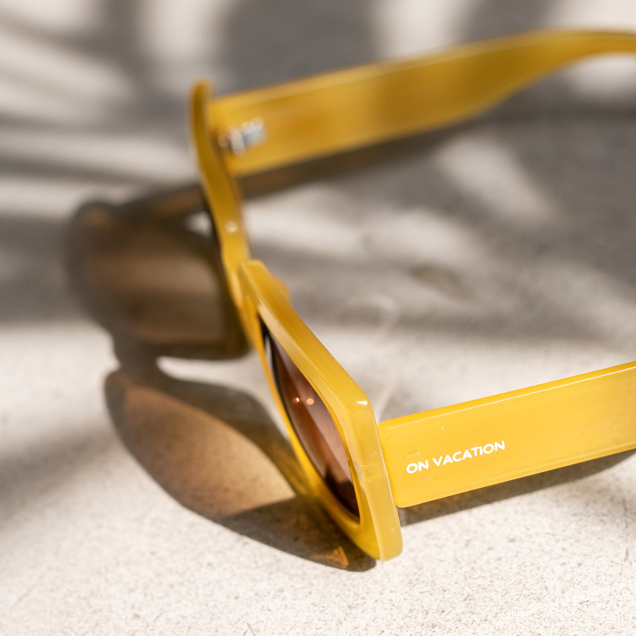 Gelbe Sonnenbrille mit der Aufschrift "ON VACATION" auf einem hellen Untergrund mit Schatten von Blättern.
