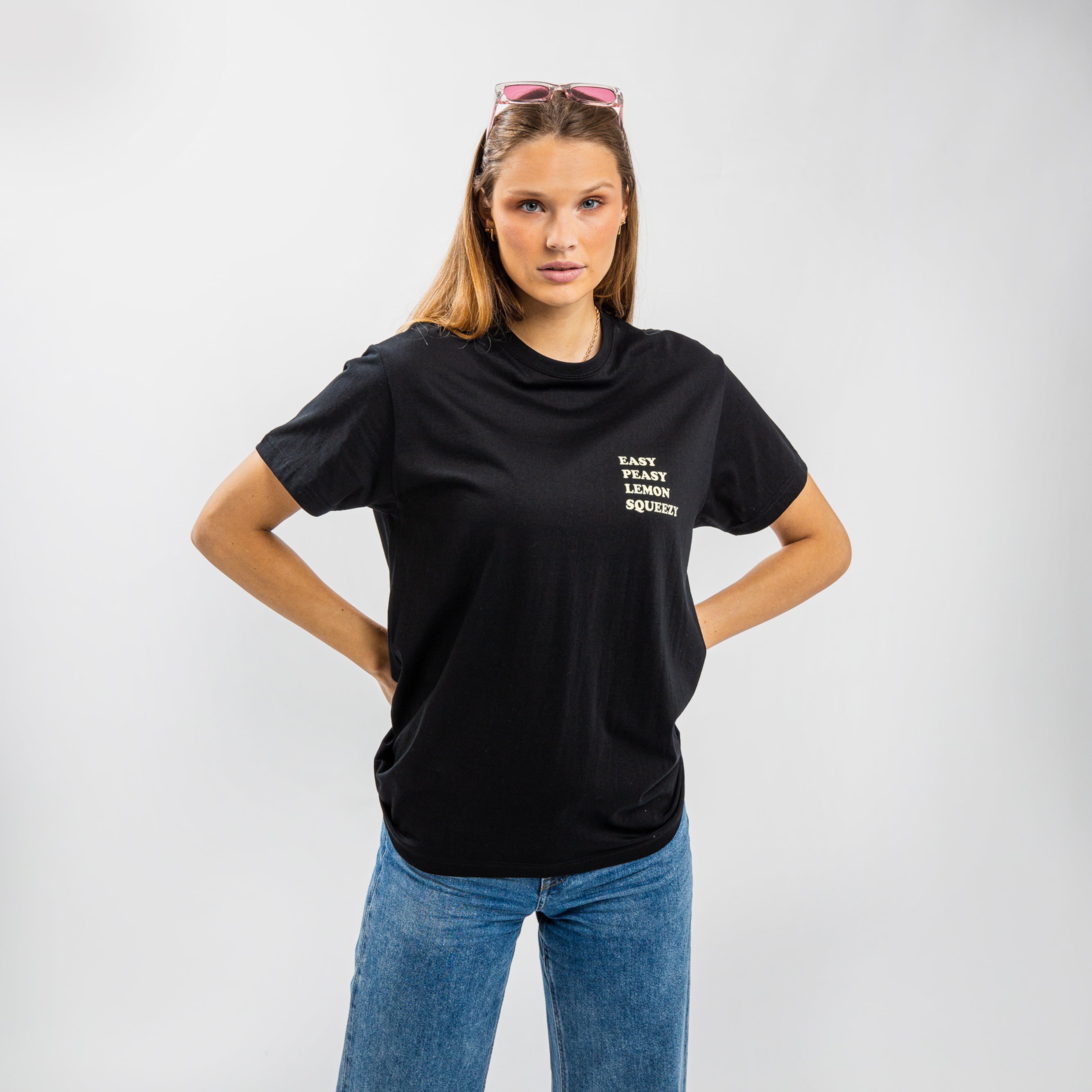 Frau in schwarzem T-Shirt mit dem Aufdruck "EASY PEASY LEMON SQUEEZY" und blauer Jeans, posiert vor hellem Hintergrund.