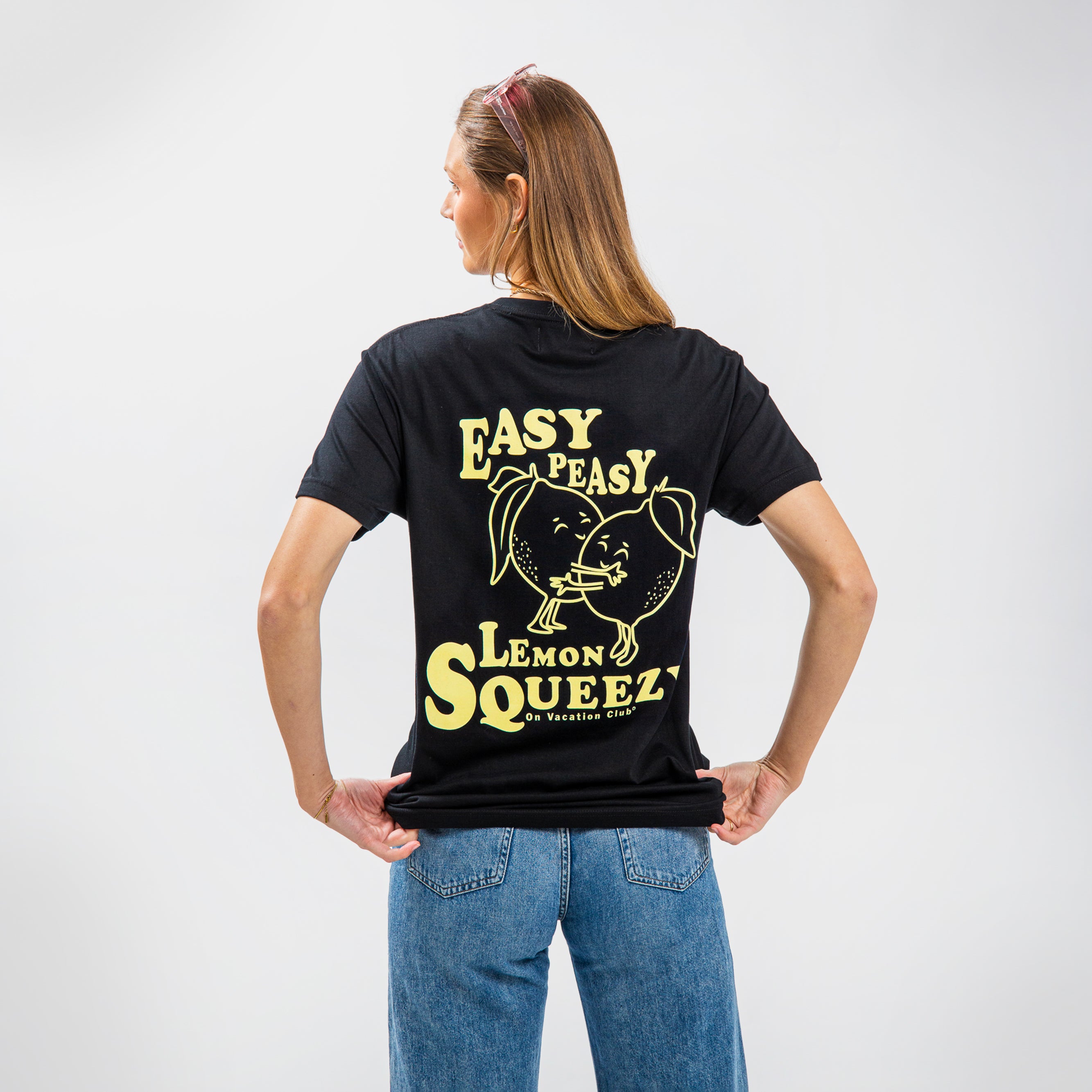 Frau in Jeans und schwarzem T-Shirt mit gelbem Aufdruck "EASY PEASY LEMON SQUEEZY" und Zitronenillustration.