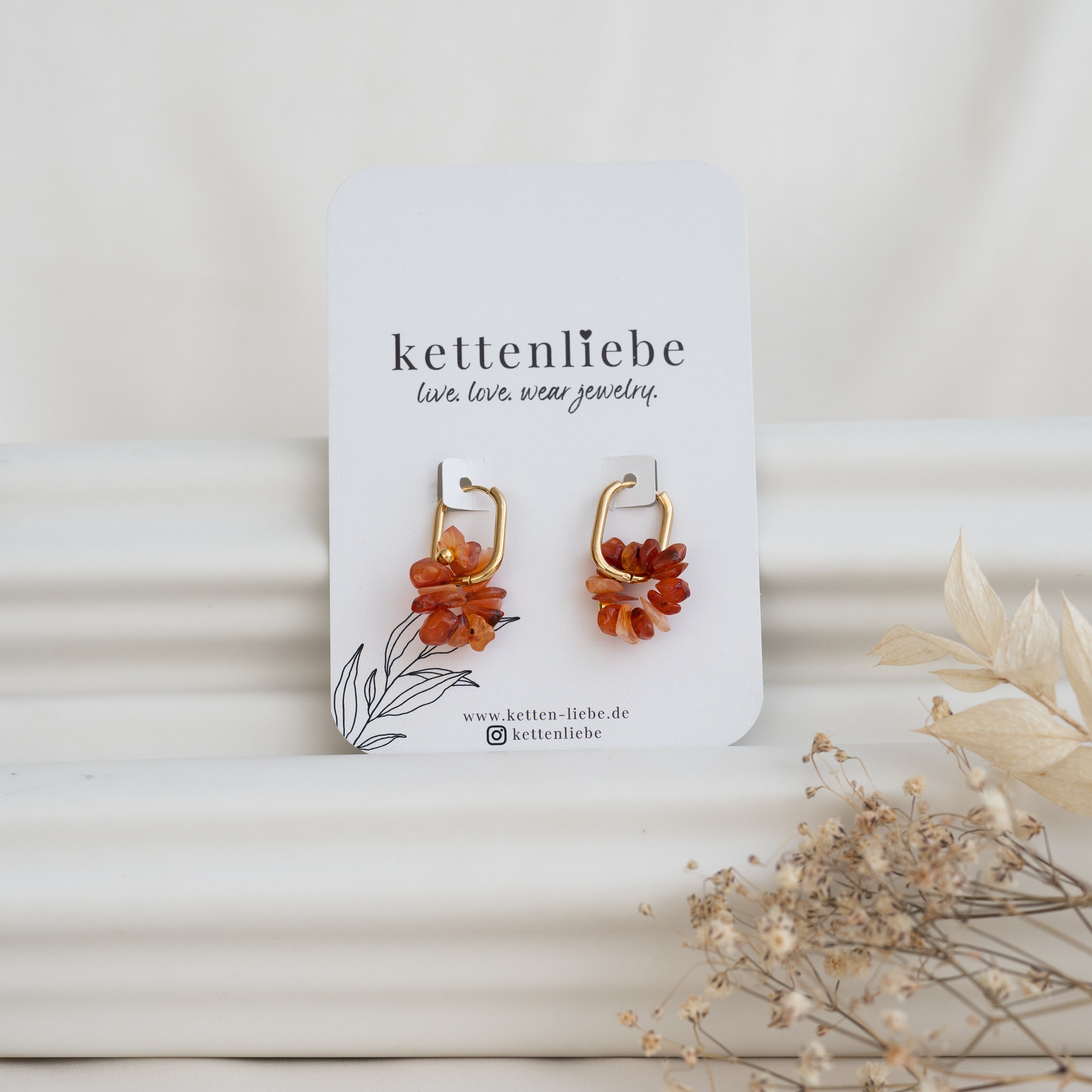Ein Paar goldene Ohrringe mit orangefarbenen Blumenakzenten auf einer Marken-Kettenliebe-Karte, vor einem weißen Hintergrund mit getrockneten Blumen im Vordergrund.