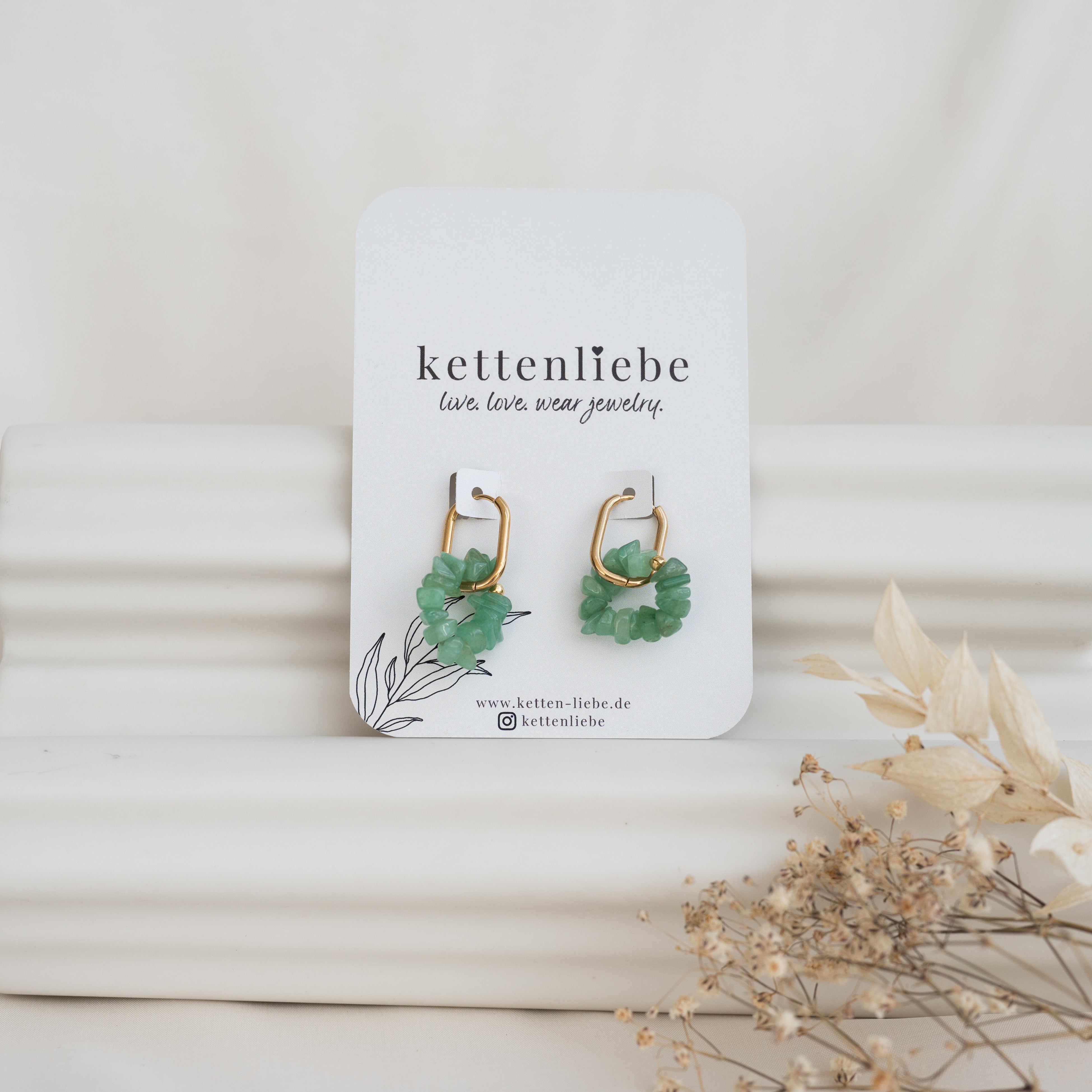 Ein Paar goldene Ohrringe mit grünen Steinen auf einer weißen Karte mit der Aufschrift "kettenliebe", vor einem weißen, strukturierten Hintergrund mit getrockneten Blumen im Vordergrund.