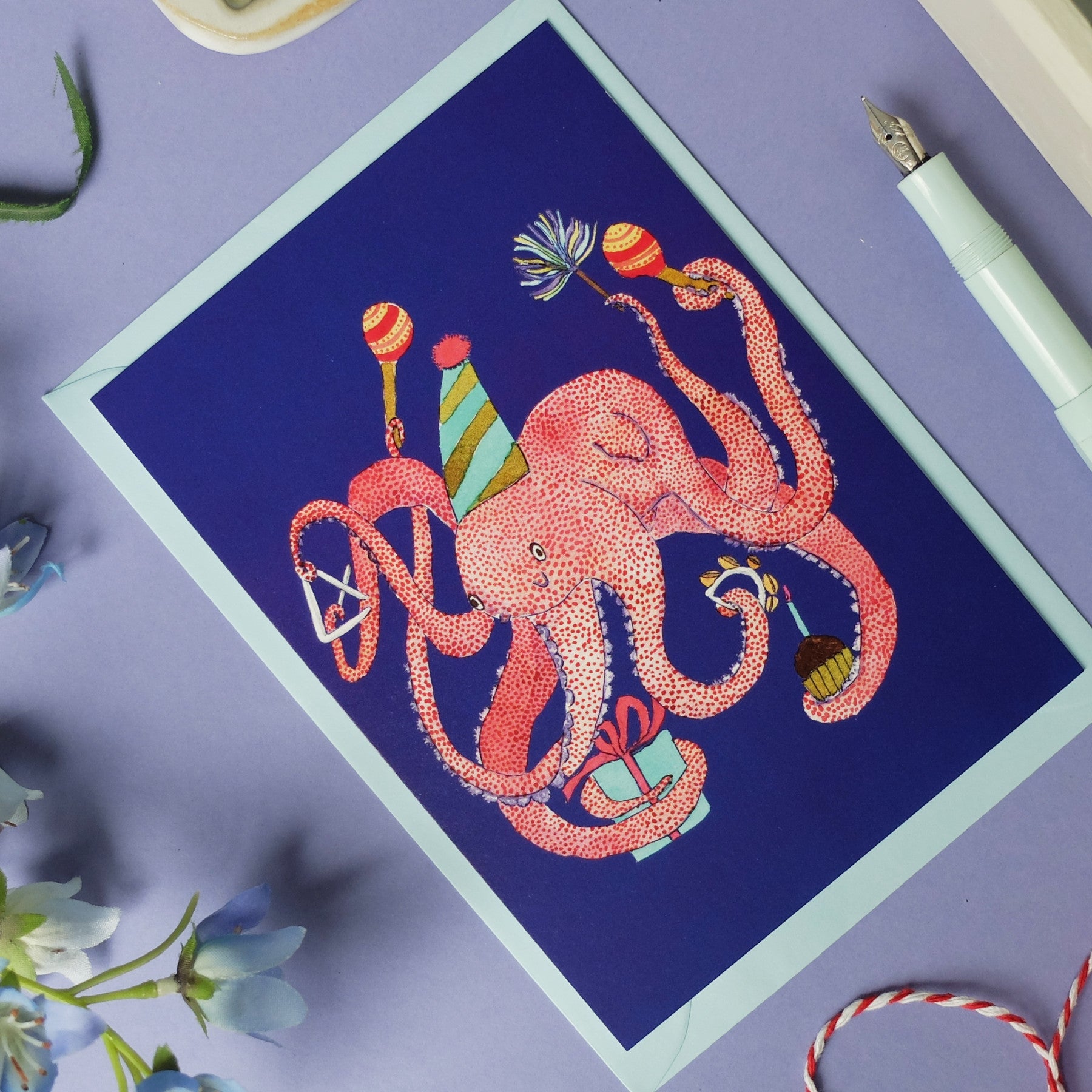 Eine Grußkarte mit einem rosa Kraken, der einen Partyhut trägt und Geburtstagsartikel wie Luftballons, ein Geschenk, einen Kuchen und ein Partyhorn auf einem blauen Hintergrund hält, umgeben von Blumen, einem Füllfederhalter und einer lila Fläche.
