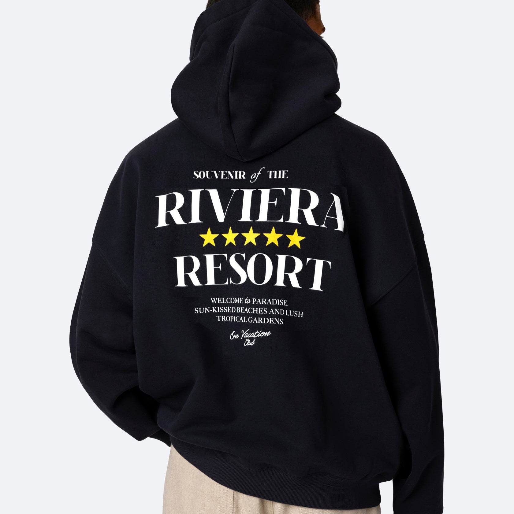 Eine Person trägt einen schwarzen Kapuzenpulli mit weißer Schrift und fünf gelben Sternen auf dem Rücken, auf dem "Souvenir of the Riviera Resort" steht und darunter ein kleinerer Aufdruck zu sehen ist. Die Person blickt von der Kamera weg.