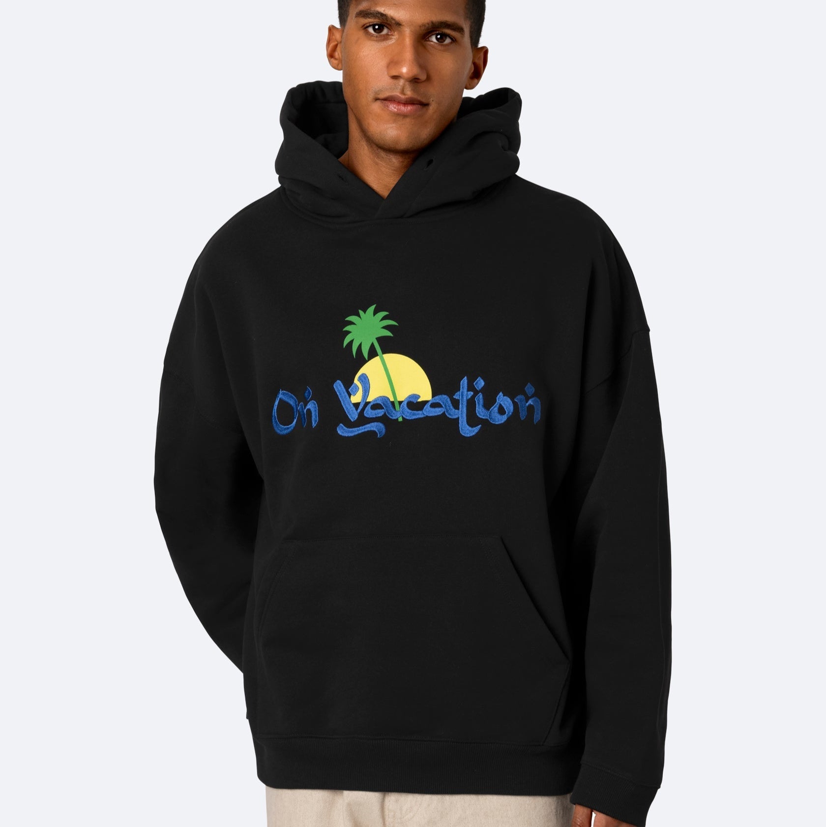 Eine Person trägt einen schwarzen Kapuzenpulli mit einer bunten Grafik einer Palme, Sonne und blauem Text, der On Vacation lautet. Sie stehen vor einem einfachen hellen Hintergrund.