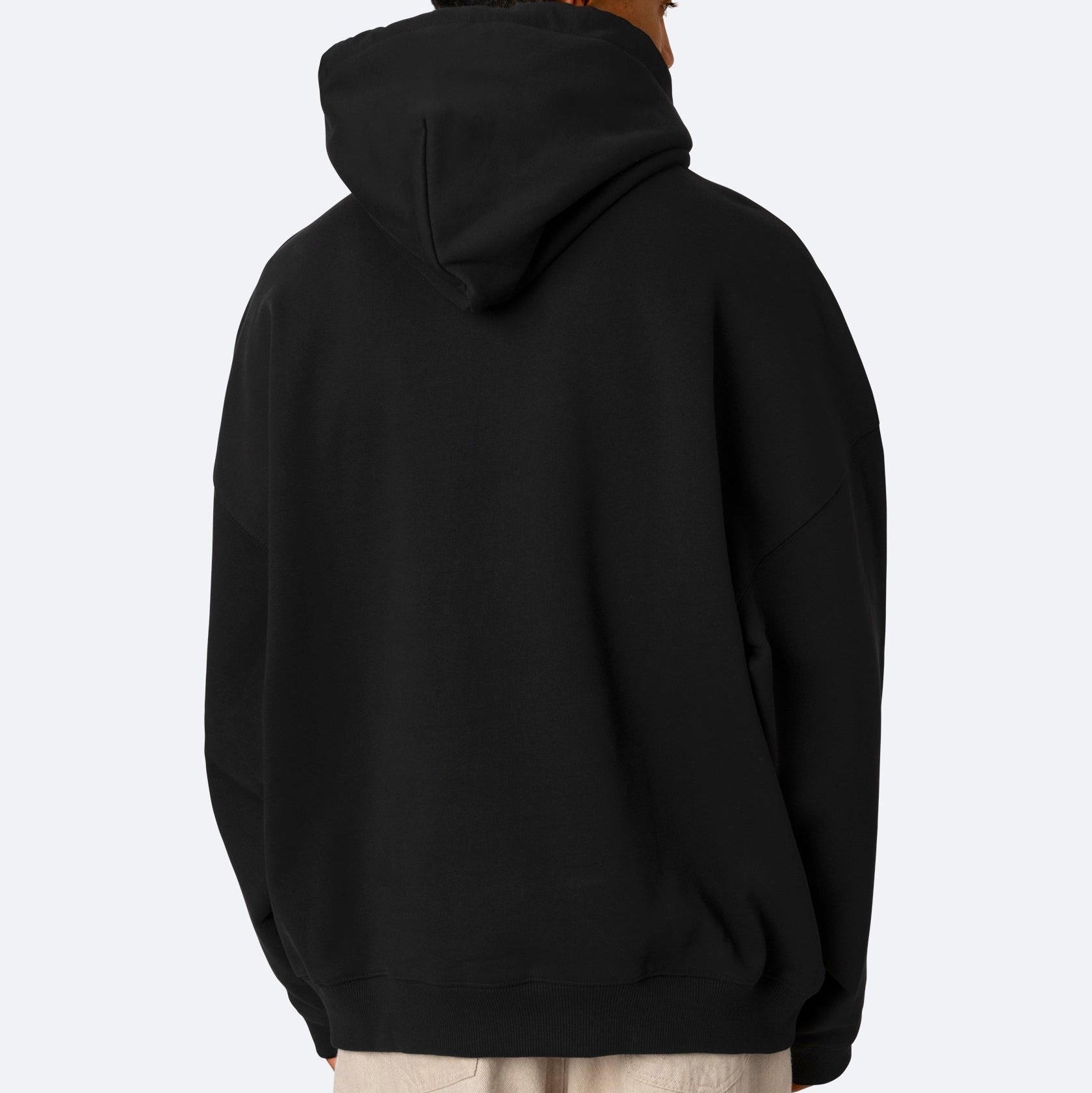 Eine Person wird von hinten gezeigt, die einen weiten schwarzen Kapuzenpullover mit aufgesetzter Kapuze und eine helle Hose trägt und vor einem einfarbigen hellen Hintergrund steht.