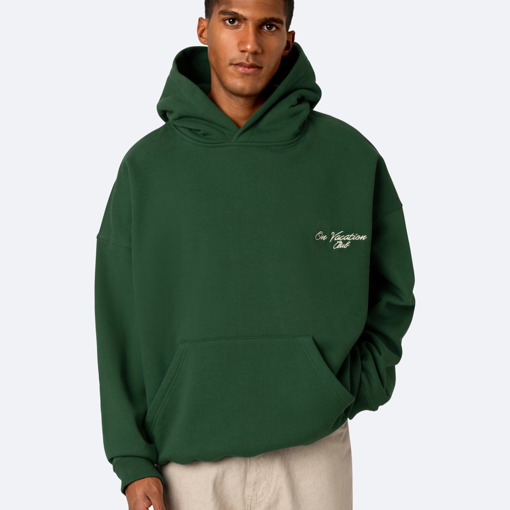 Eine Person, die einen dunkelgrünen Kapuzenpullover mit der Aufschrift On Vacation Club auf der linken oberen Brustseite trägt, steht mit einer Hand in der Tasche vor einem schlichten hellen Hintergrund.