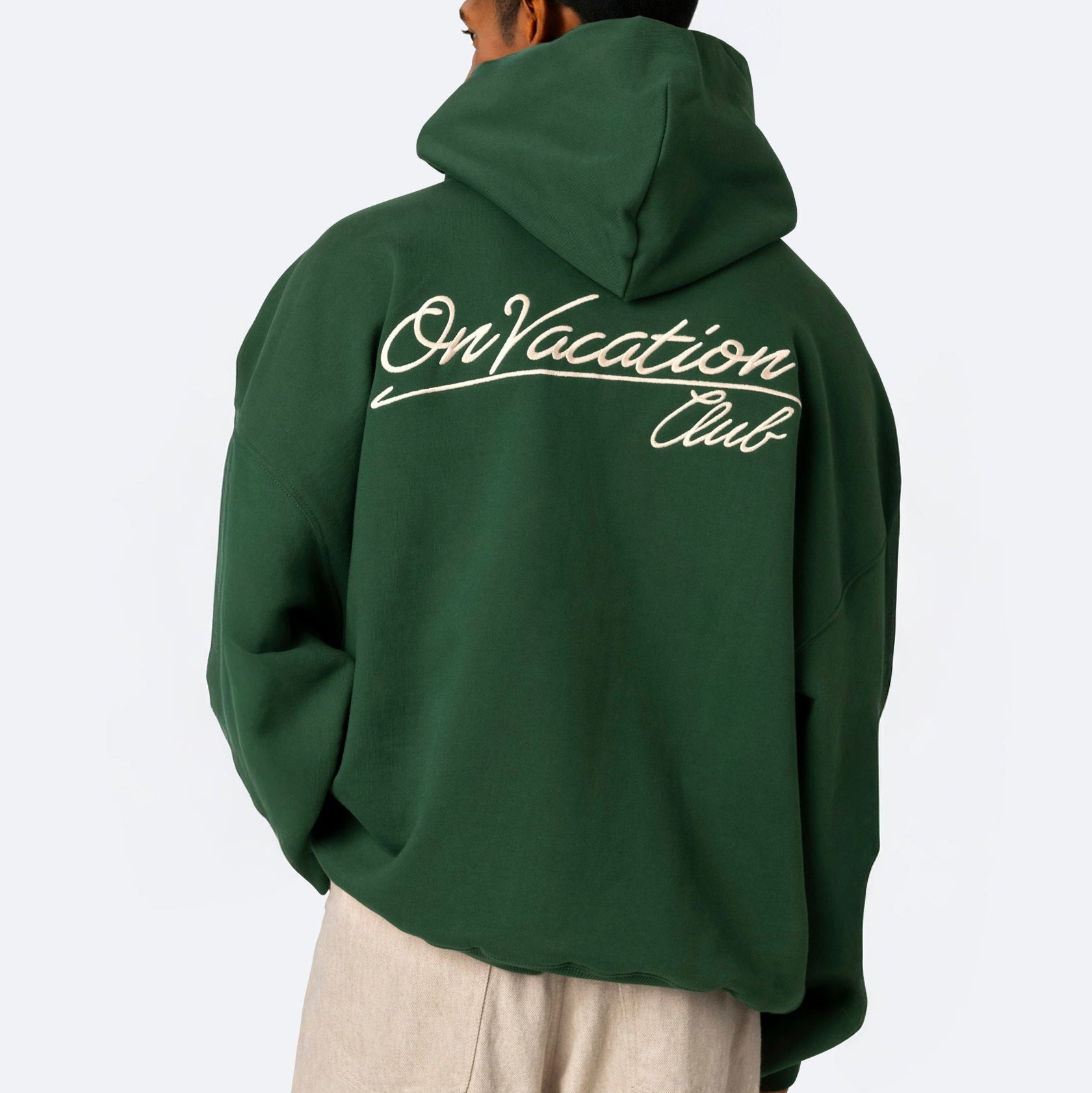 Eine Person, die ein grünes Kapuzensweatshirt mit der Aufschrift "On Vacation Club" in weißer Schrift auf dem Rücken und eine helle Hose trägt und von der Kamera wegschaut.