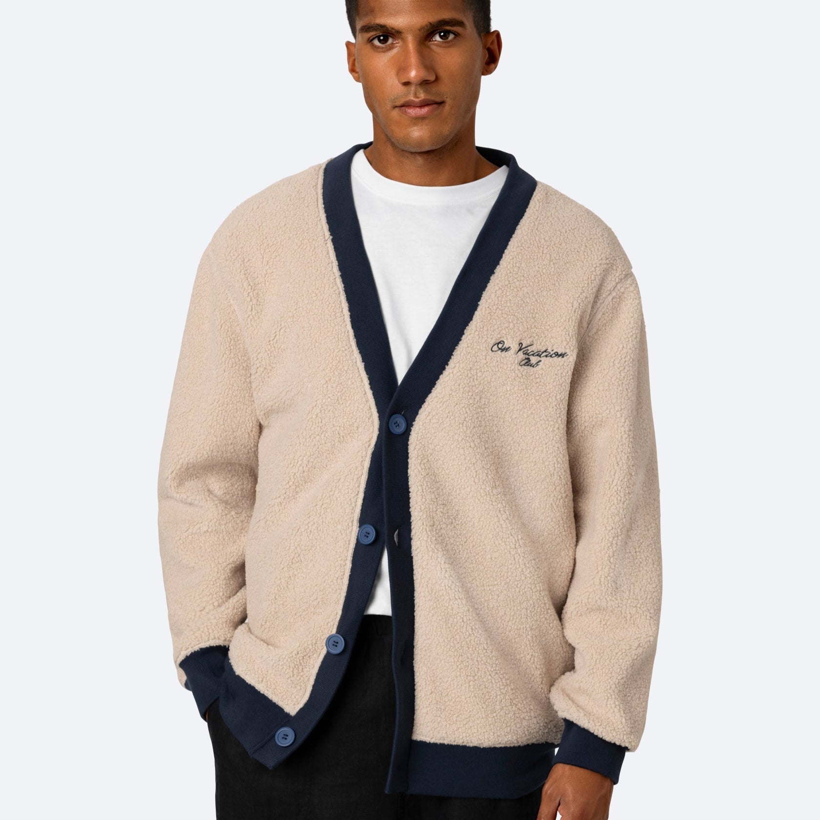 Ein Mann steht mit dem Gesicht nach vorne und trägt eine beigefarbene Fleecejacke mit marineblauem Saum über einem weißen T-Shirt und einer schwarzen Hose. Die Strickjacke hat Knöpfe und ein gesticktes Logo auf der Brust. Der Hintergrund ist einfarbig hellgrau.