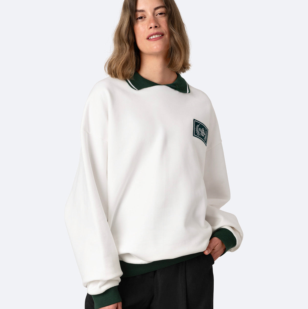 Eine Person mit schulterlangen braunen Haaren trägt ein weißes Sweatshirt mit dunkelgrünem Kragen und Manschetten sowie eine schwarze Hose. Sie steht vor einem schlichten hellen Hintergrund und lächelt leicht.