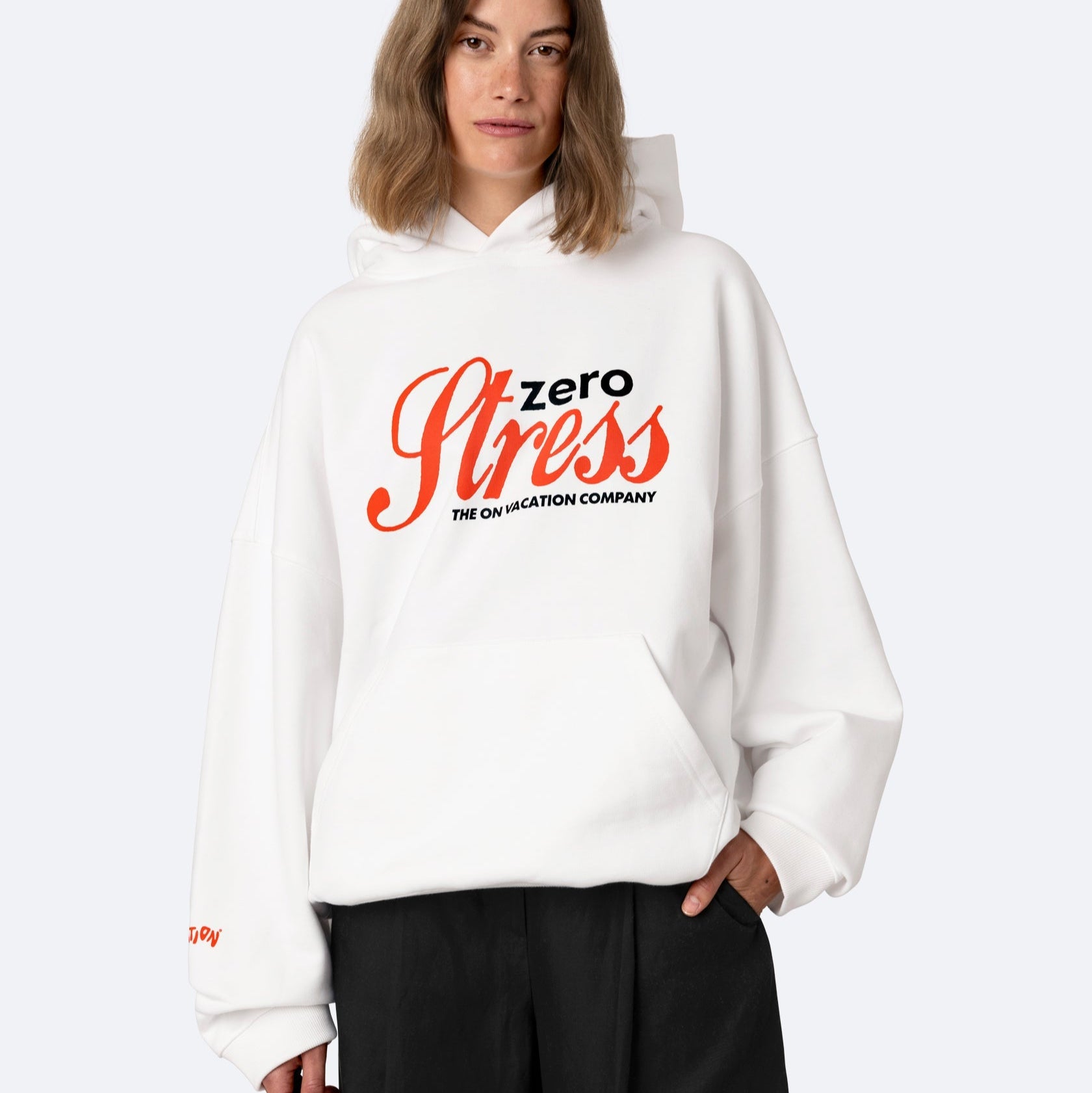 Eine Person, die einen weißen Kapuzenpulli mit der Aufschrift "Zero Stress The On Vacation Company" in fetter roter und schwarzer Schrift trägt, steht mit einer Hand in der Tasche und schaut in die Kamera.