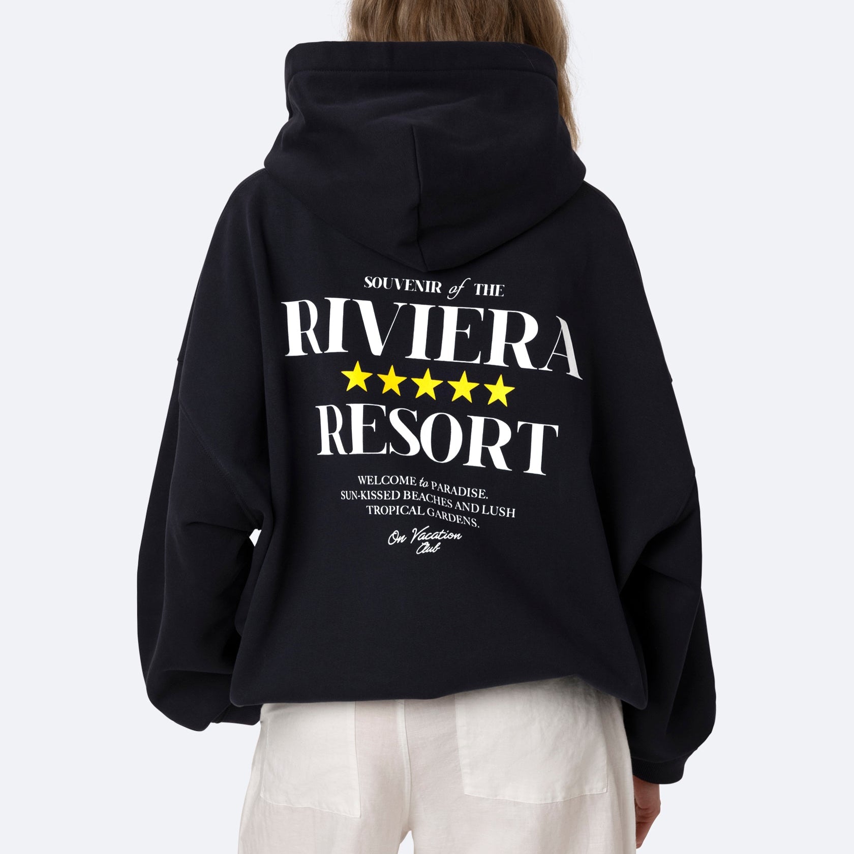 Eine Person mit langen braunen Haaren trägt einen schwarzen Kapuzenpulli mit der Aufschrift "SOUVENIR OF THE RIVIERA RESORT" und fünf gelben Sternen auf dem Rücken, gepaart mit einer weißen Hose, und blickt von der Kamera weg auf einen einfarbigen Hintergrund.