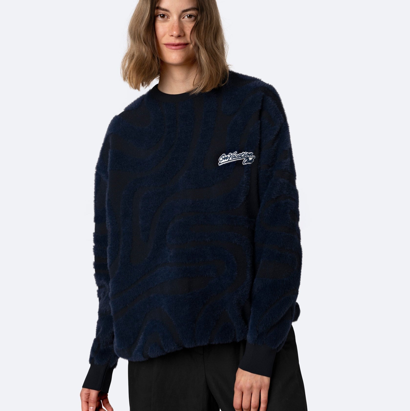 Eine Person mit schulterlangen braunen Haaren trägt einen marineblauen strukturierten Pullover mit einem abstrakten Muster und eine schwarze Hose vor einem schlichten hellen Hintergrund.