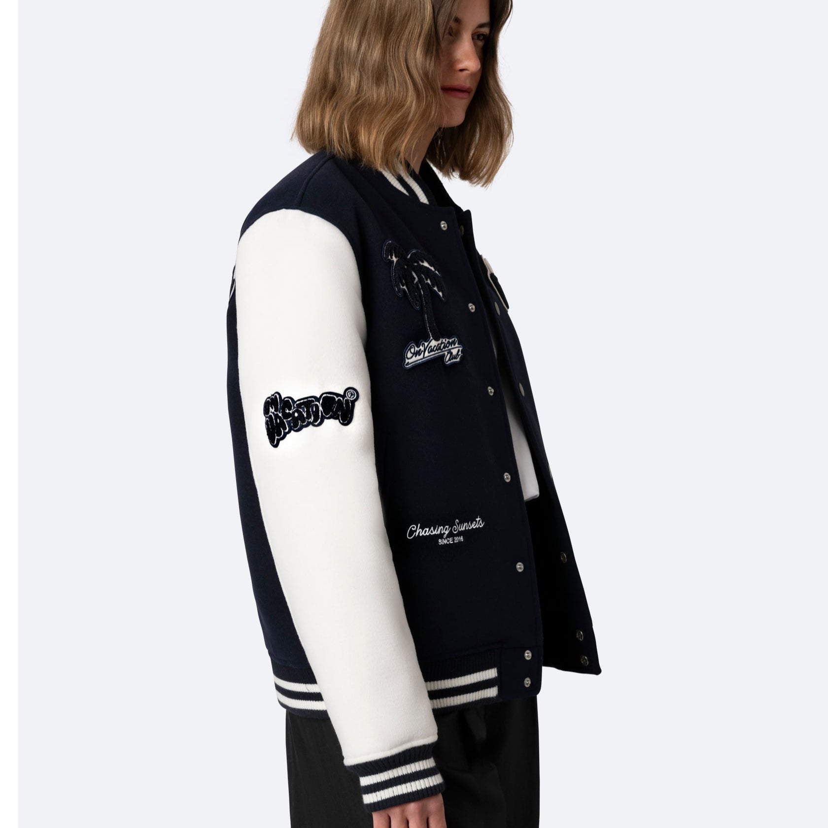 Eine Frau mit schulterlangen braunen Haaren trägt eine marineblaue Sportjacke mit weißen Ärmeln, gestreiften Manschetten und mehreren Aufnähern vor einem schlichten hellen Hintergrund.