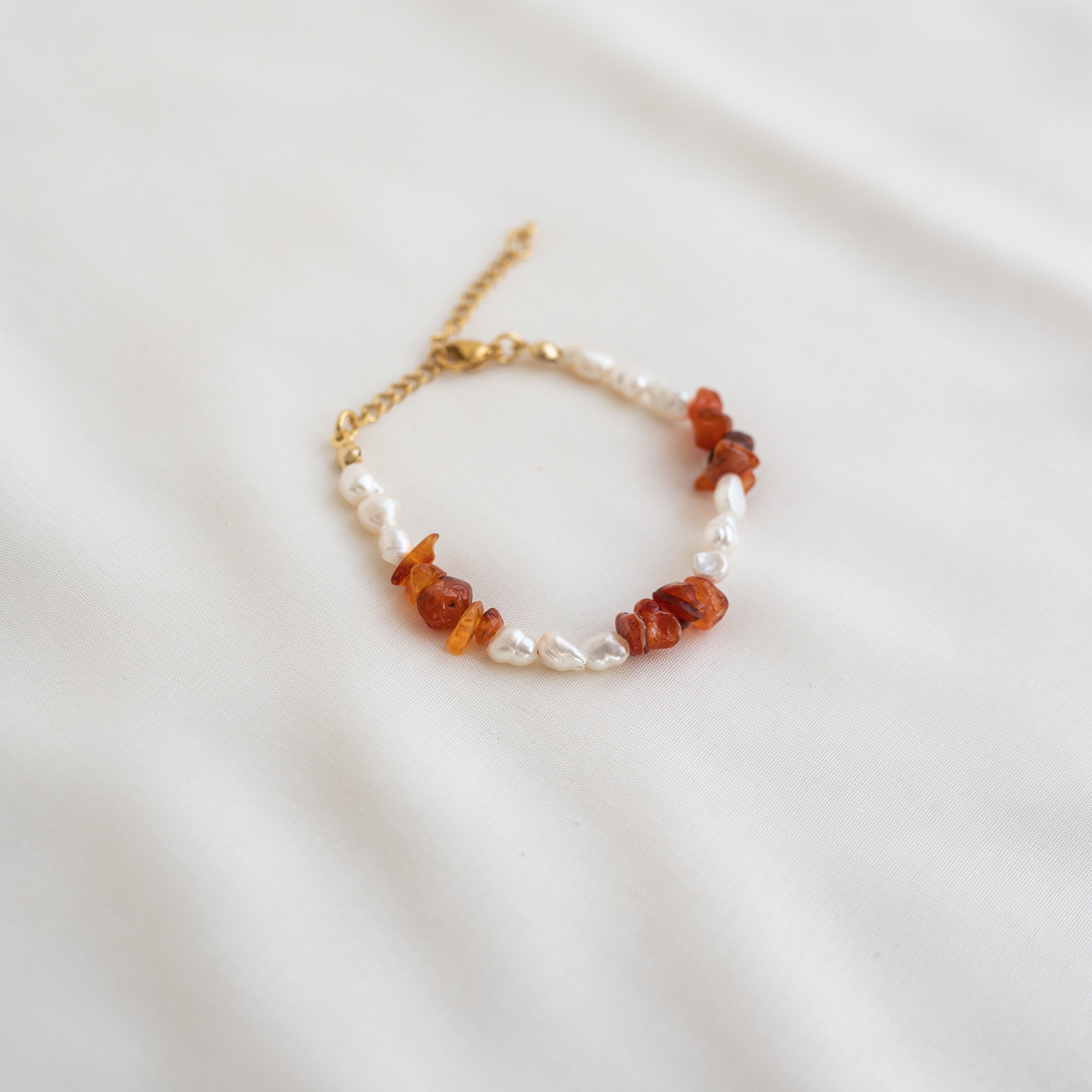 Ein Perlenarmband mit abwechselnd weißen Perlen und Bernsteinen, mit einem goldenen Kettenverschluss, auf einem weichen weißen Stoffhintergrund.