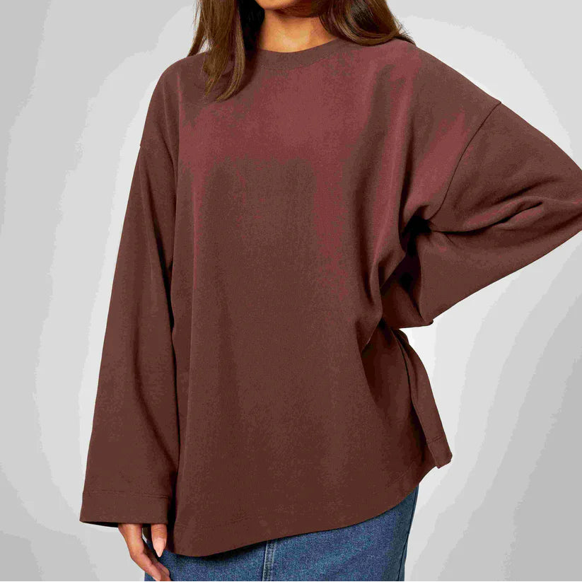 Eine Person, die ein locker sitzendes, langärmeliges braunes Oberteil und blaue Jeans trägt, steht vor einem schlichten hellgrauen Hintergrund. Der Kopf der Person ist teilweise außerhalb des Rahmens.