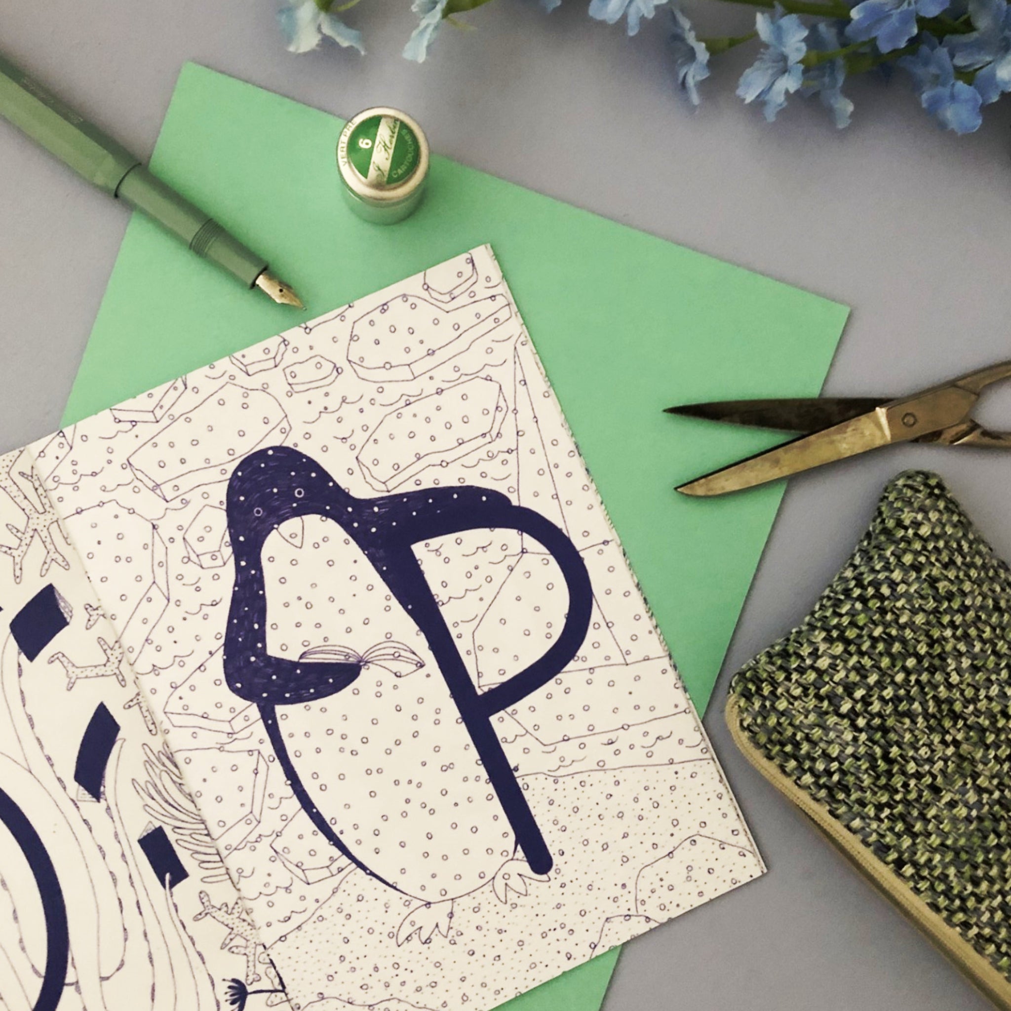 Zeichnung eines Pinguins auf einem gemusterten Papier, umgeben von Stift, Schere, grüner Karte und Häkelarbeit.