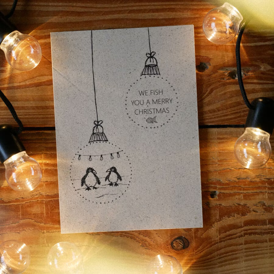 Weihnachtskarte mit zwei gezeichneten Pinguinen und dem Text "We fish you a Merry Christmas", umgeben von Lichterketten.