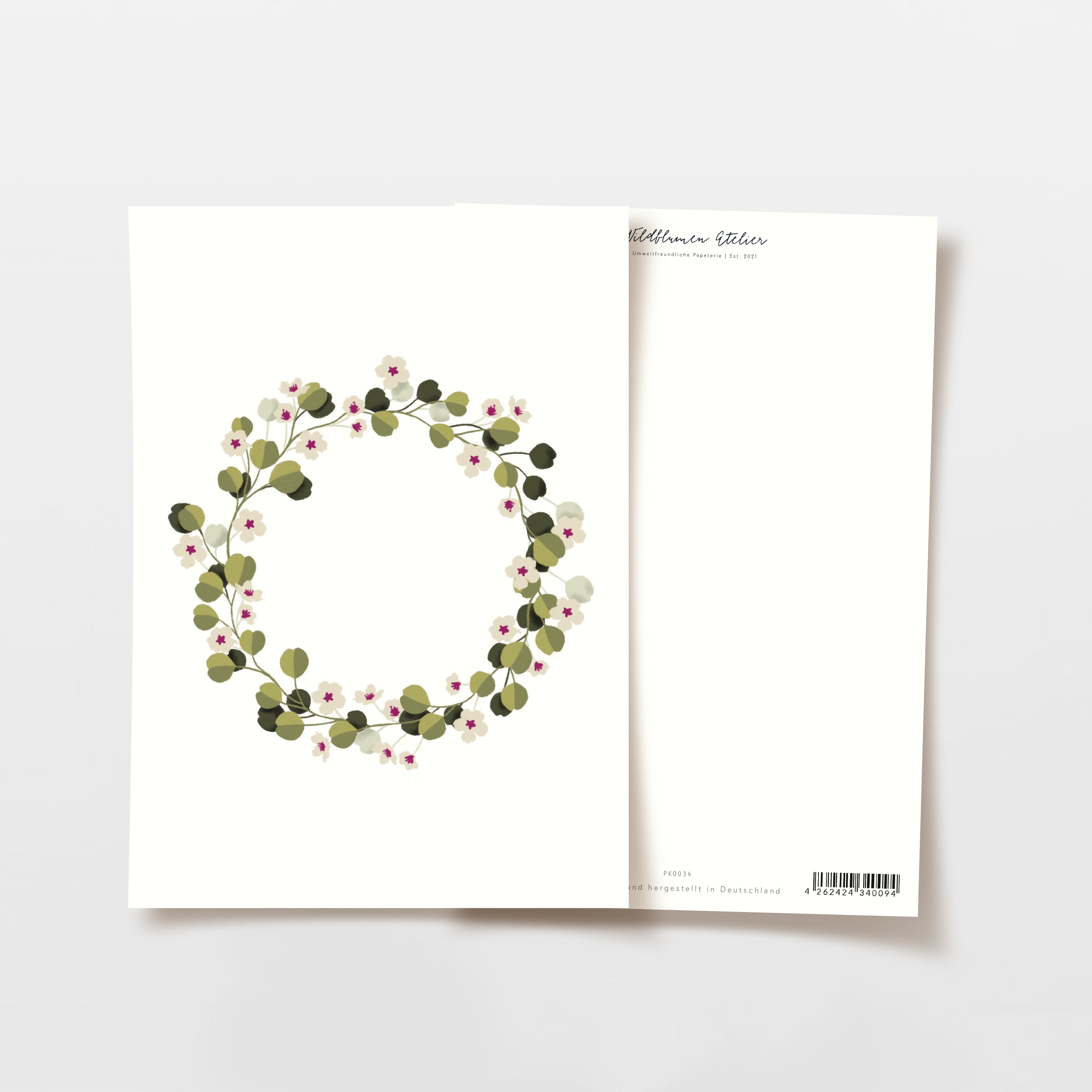 Zwei Grußkarten auf einer weißen Fläche. Die vordere Karte zeigt einen kreisförmigen Kranz aus grünen Blättern und kleinen rosa und weißen Blumen. Die Rückseite ist größtenteils leer, mit einem kleinen Text und einem Strichcode am unteren Rand.