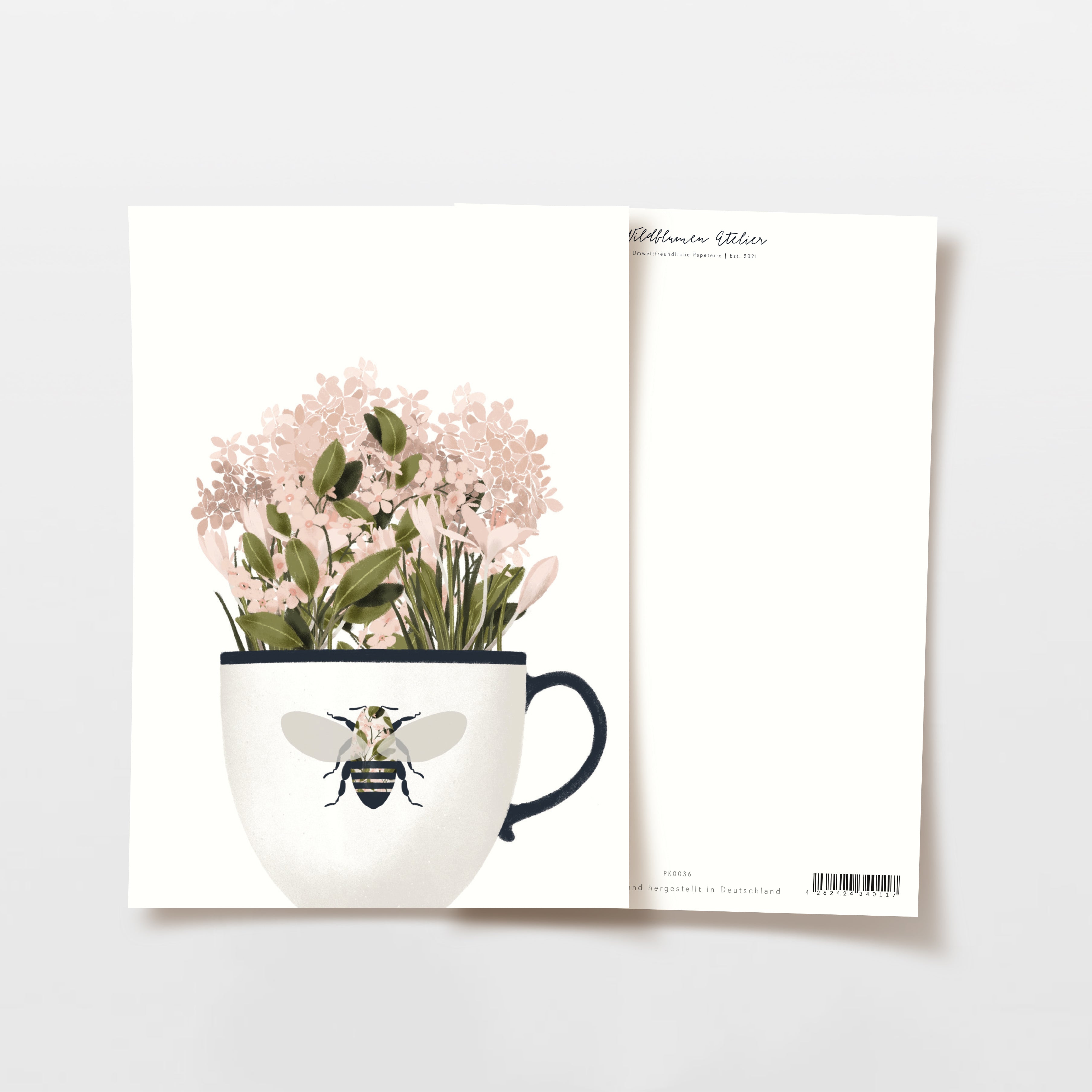 Eine Grußkarte mit einer Illustration von rosa Blumen in einer weißen Teetasse mit einem Bienenmotiv auf der Vorderseite, dargestellt mit der Vorder- und Rückseite der Karte nebeneinander auf einem weißen Hintergrund.