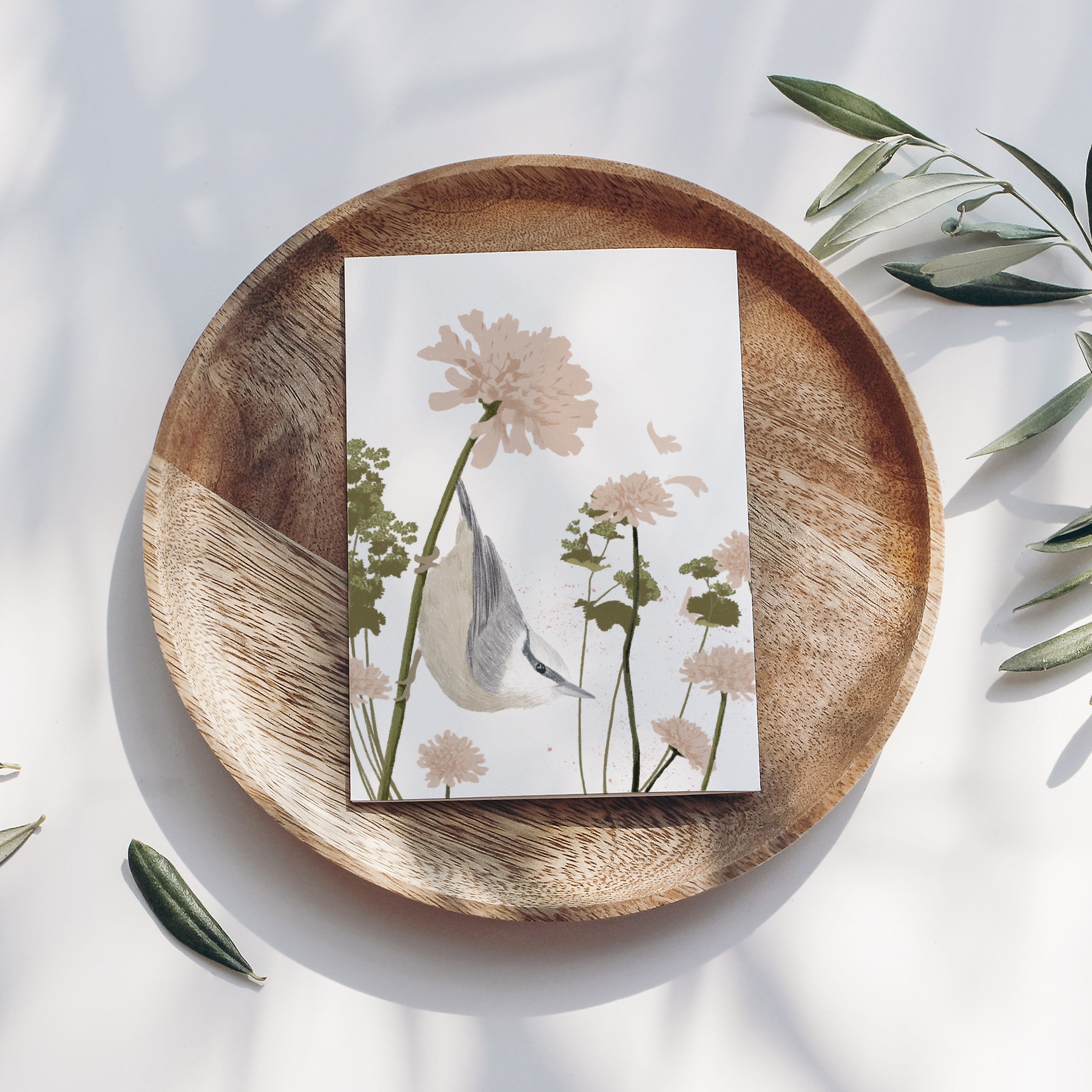 Holzteller mit einer Karte, die Blumen und einen Vogel zeigt, daneben Olivenzweige auf weißem Hintergrund.