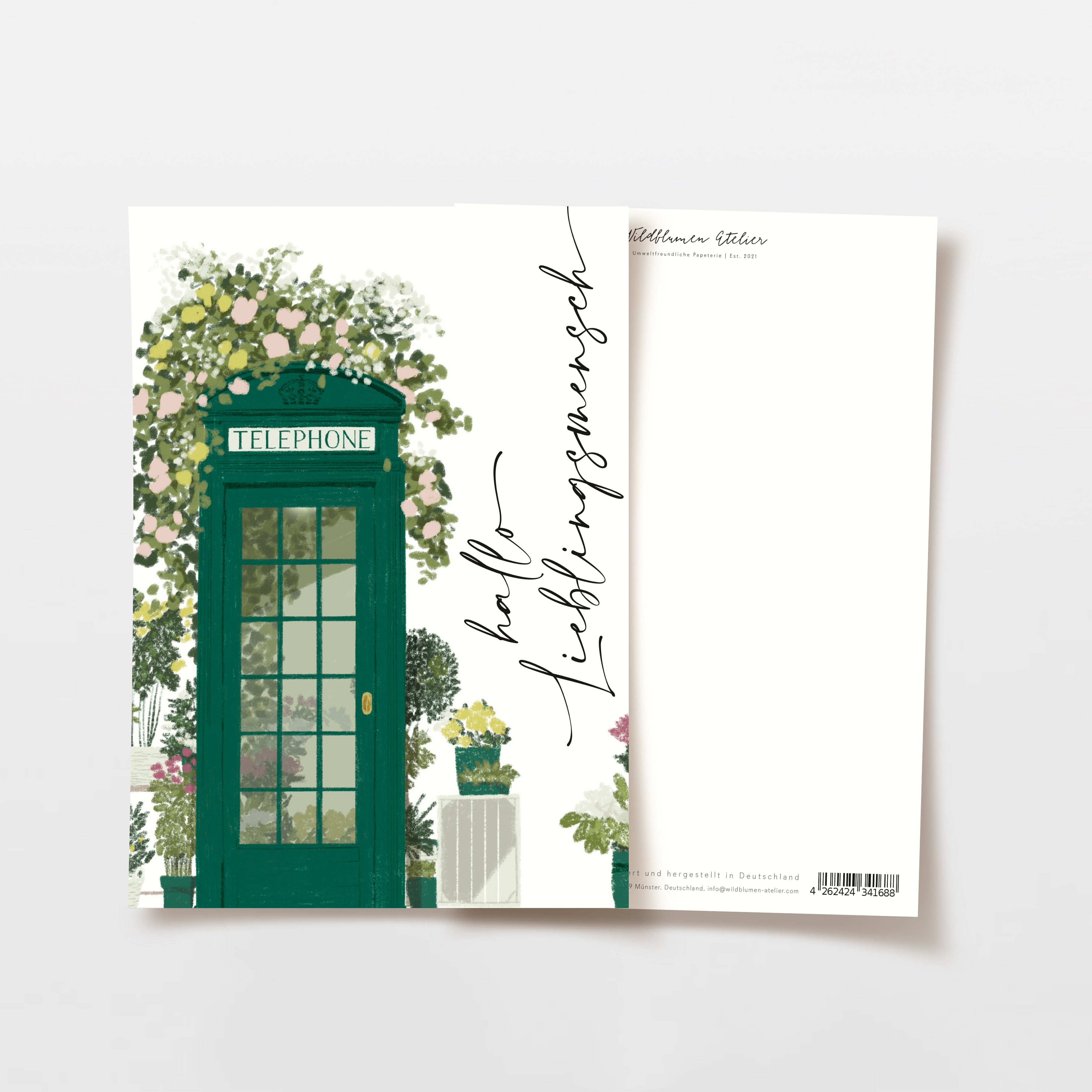 Eine Postkarte mit einer grünen Telefonzelle, umgeben von blühenden Blumen und Pflanzen. Auf der Vorderseite steht "Hallo Lieblingsmensch" in Schreibschrift, die Rückseite ist frei zum Beschreiben.