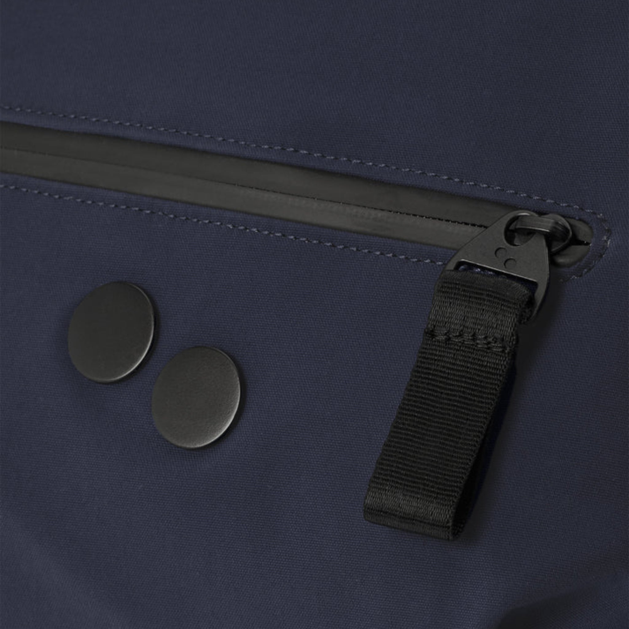 Nahaufnahme einer marineblauen Stoffoberfläche mit einem schwarzen Reißverschluss mit einer Zuglasche aus Stoff und zwei runden schwarzen Knöpfen daneben.