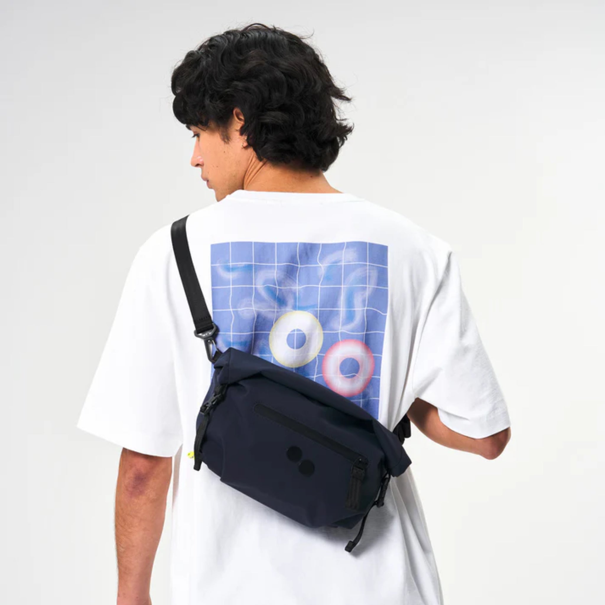 Eine Person mit dunklem, gewelltem Haar trägt ein weißes T-Shirt mit einer Grafik aus zwei pastellfarbenen Ringen auf einem blauen Gitterhintergrund. Sie hat eine marineblaue Umhängetasche über die Schulter gehängt und blickt von der Kamera weg.