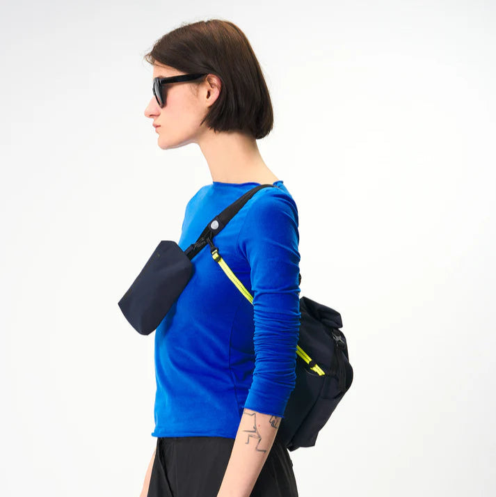 Eine Person mit kurzen braunen Haaren, die eine Sonnenbrille, ein hellblaues langärmeliges Hemd und eine schwarze Hose trägt und eine schwarze Umhängetasche mit sich führt, steht vor einem schlichten hellen Hintergrund.